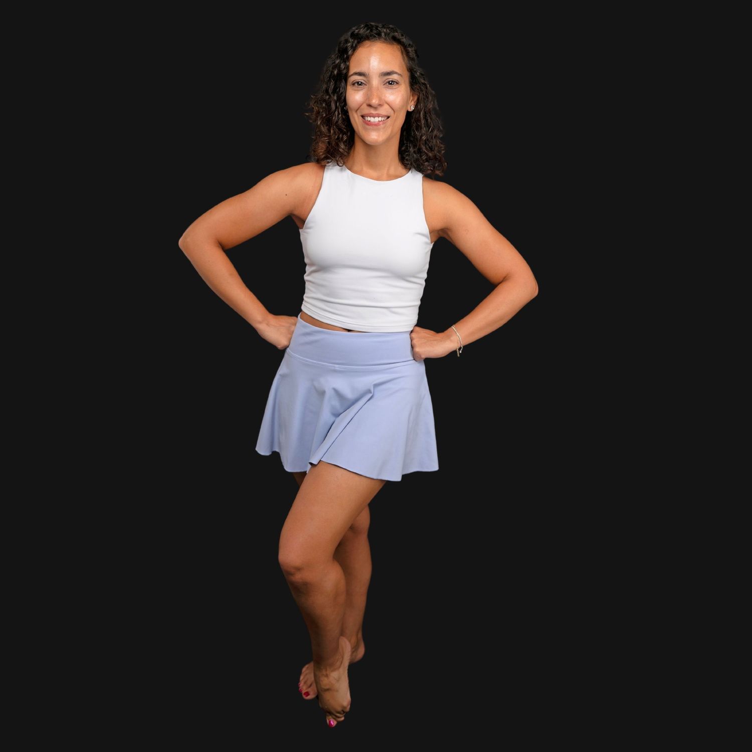 Woman posing in white crop top and light blue athletic skort against dark background.