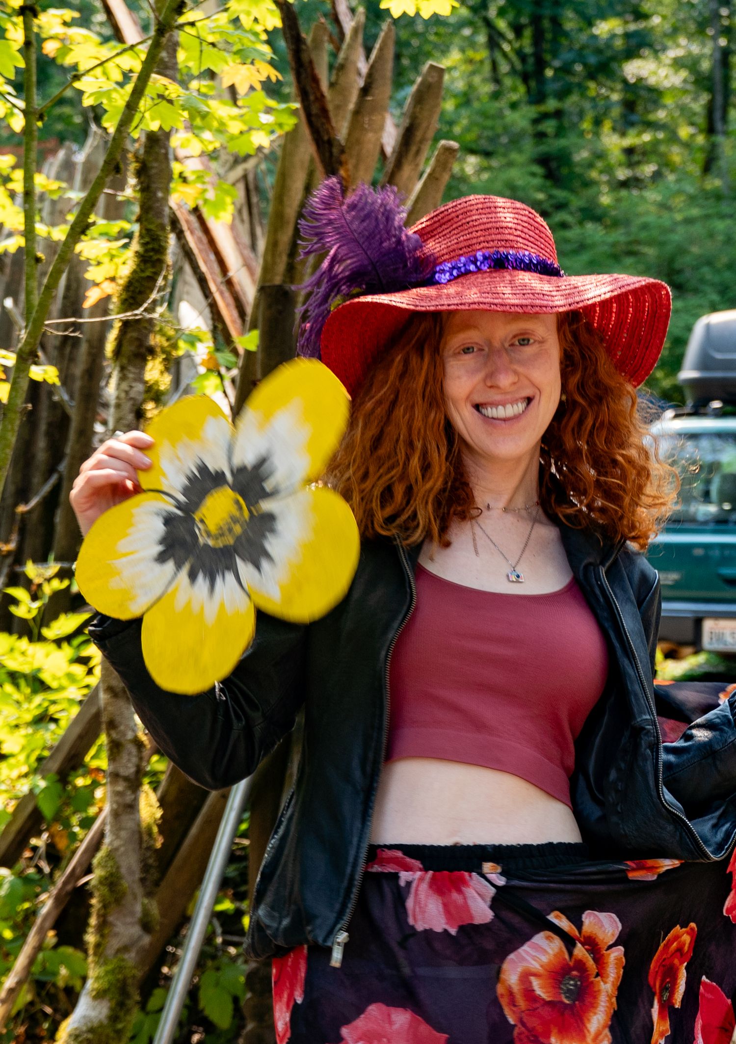 Smiling redhead woman in red wide-brim hat holding yellow flower art outdoors in forest setting.