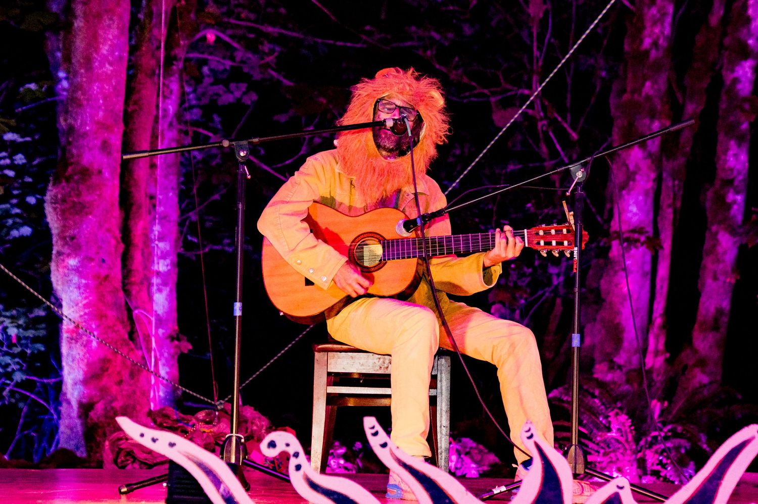 A musician with curly hair plays acoustic guitar on stage surrounded by colorful forest-themed decorations.