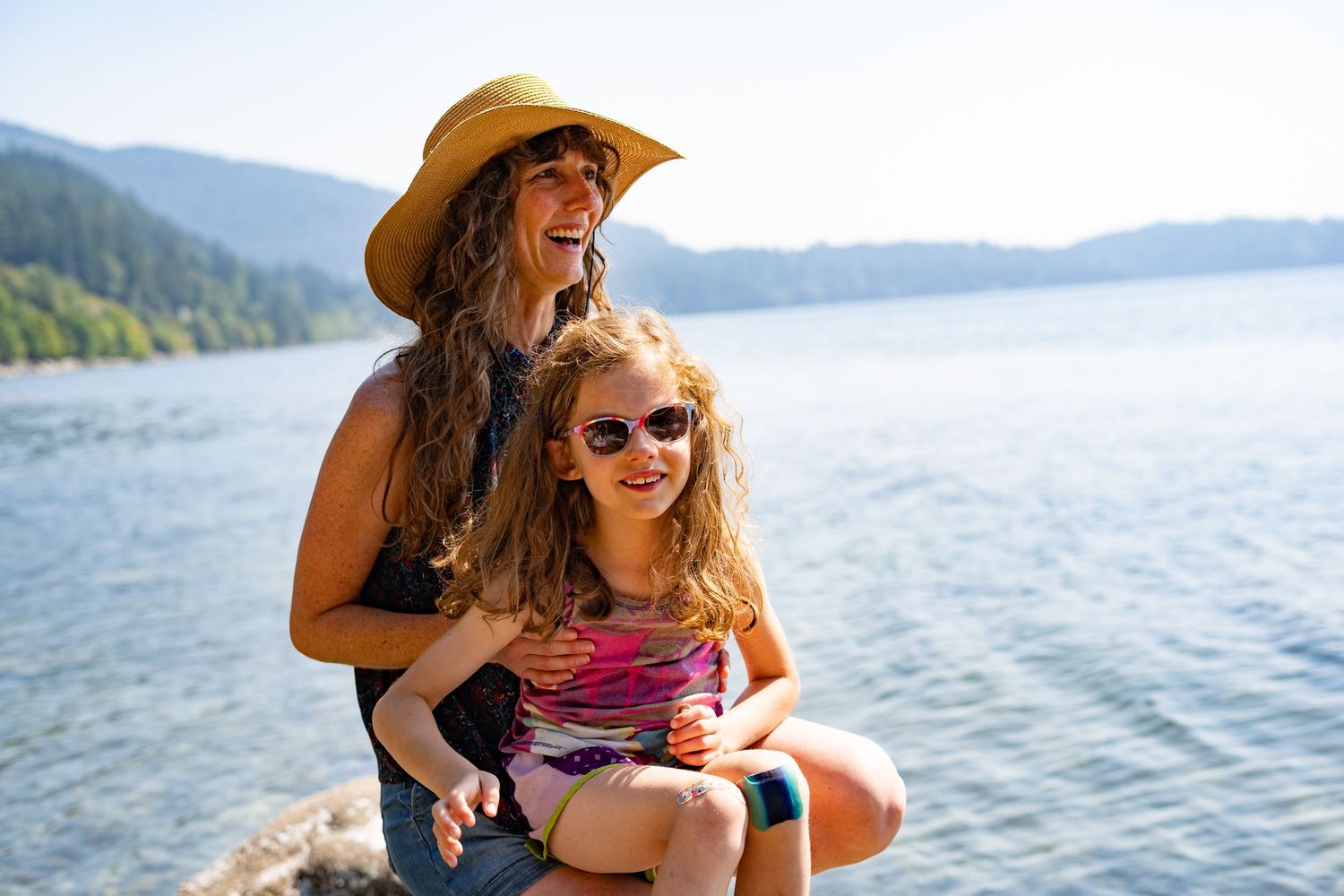Woman and young girl in sunglasses smile together on rocky lakeside with mountains and blue water behind them.