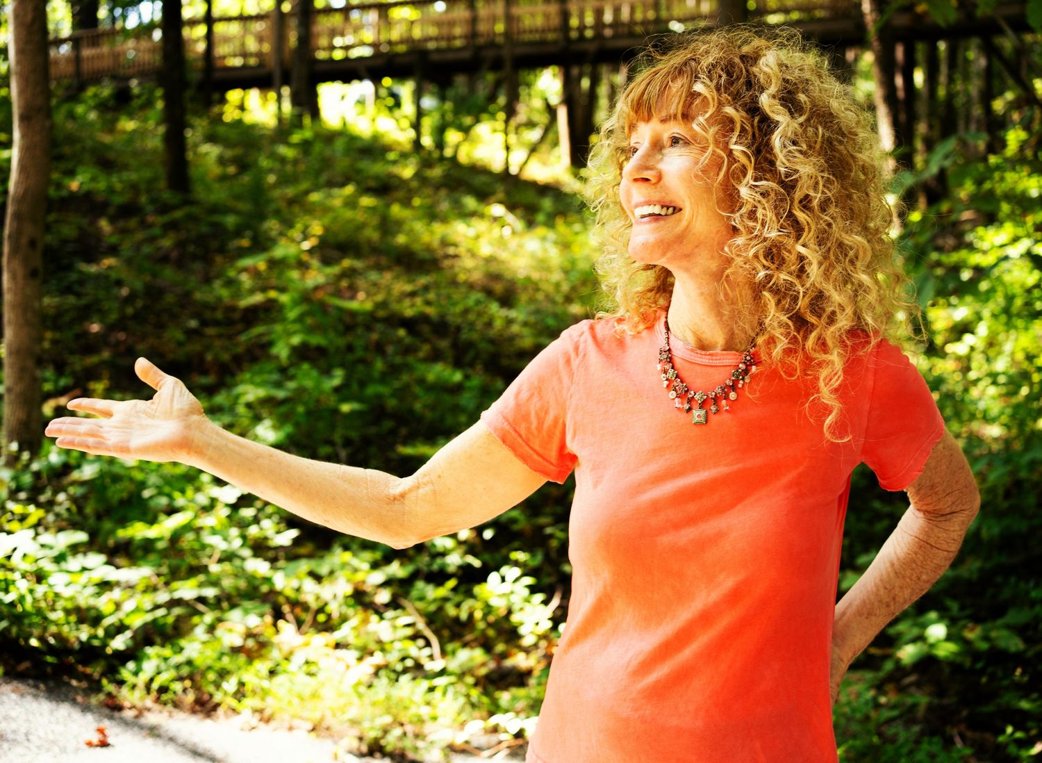 A photo of a woman with curly orange hair, a bright orange top and she is smiling and looking out into the distance.