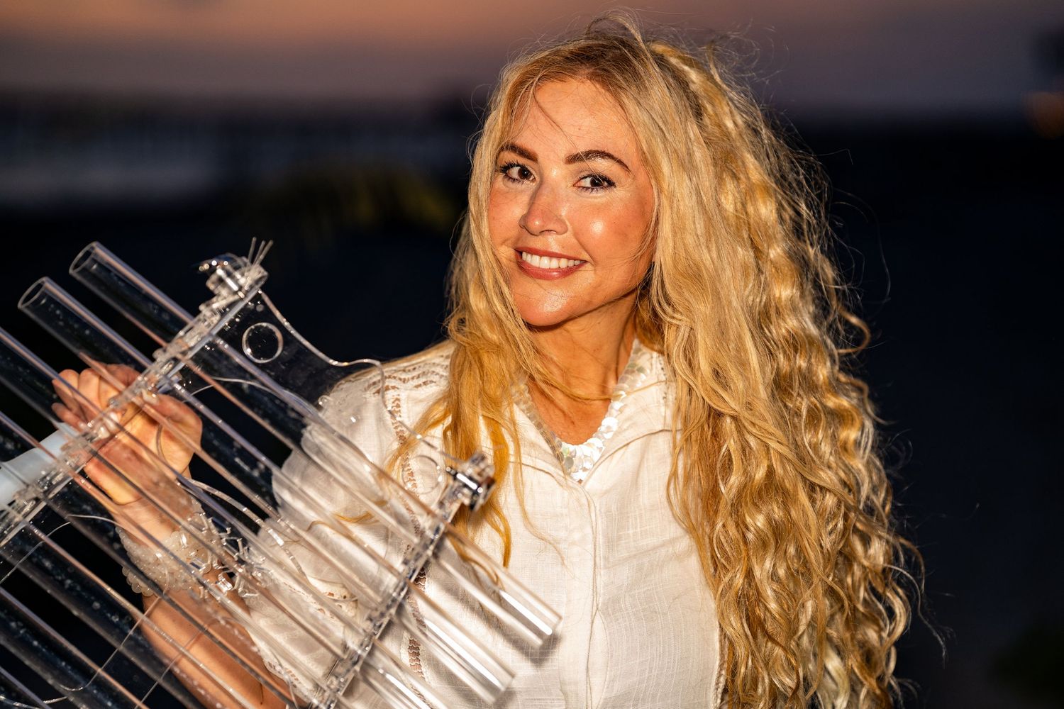 A photo of a beautiful blonde woman smiling at the camera and wearing a white button up top.
