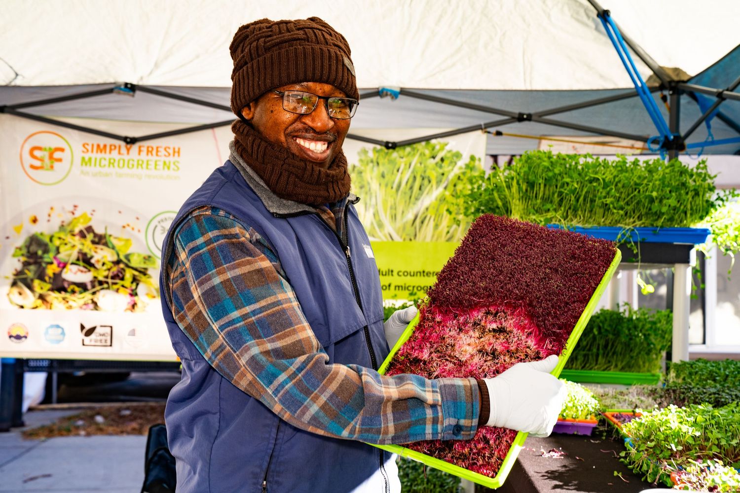 Vendor smiling and showcasing fresh purple microgreens at Boulder Farmers Market, locally grown Colorado produce.