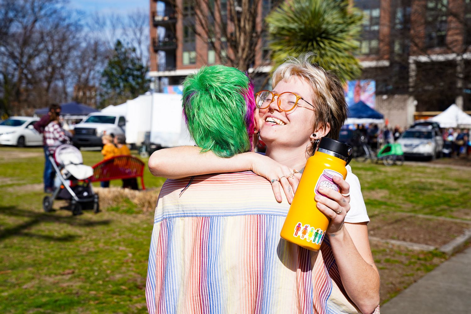 Two friends hug in downtown Boulder as one laughs with a toothpick in her mouth, wearing orange-tinted glasses and holding a yellow water.