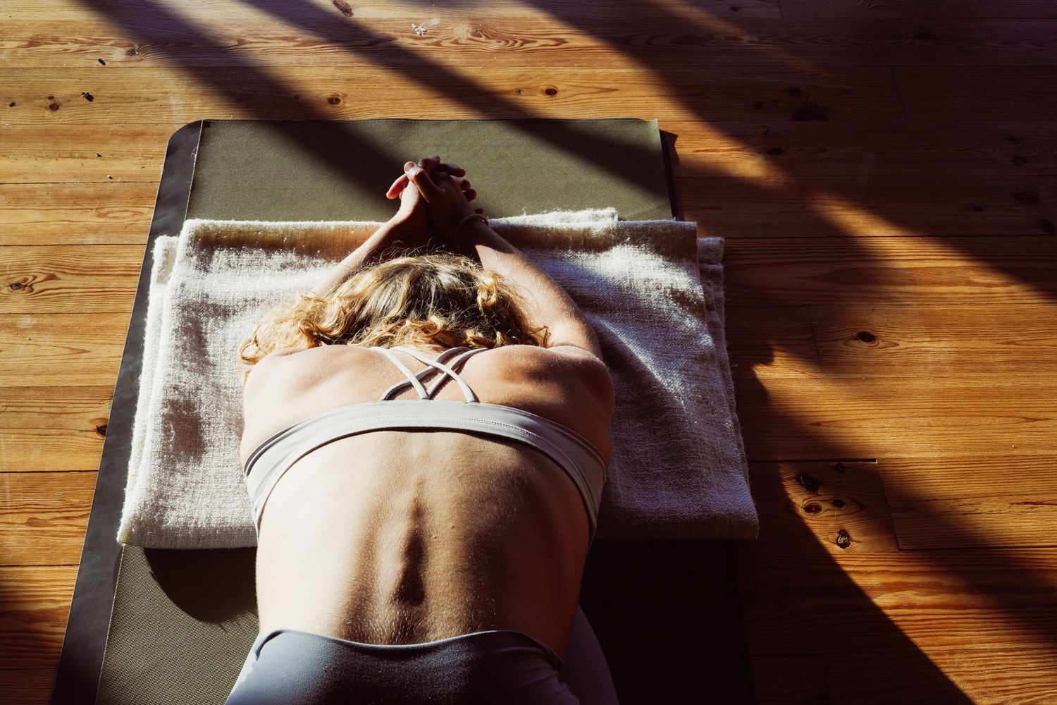 Woman lying face down on yoga mat in sunlit wooden floor studio.