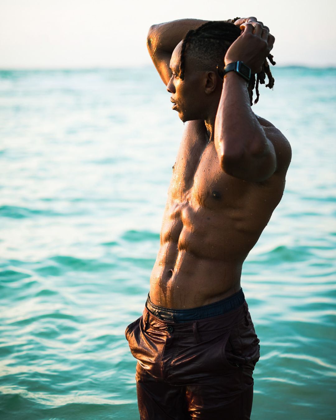 Shirtless athletic man with dreadlocks poses on a tropical beach with turquoise ocean water in the background.