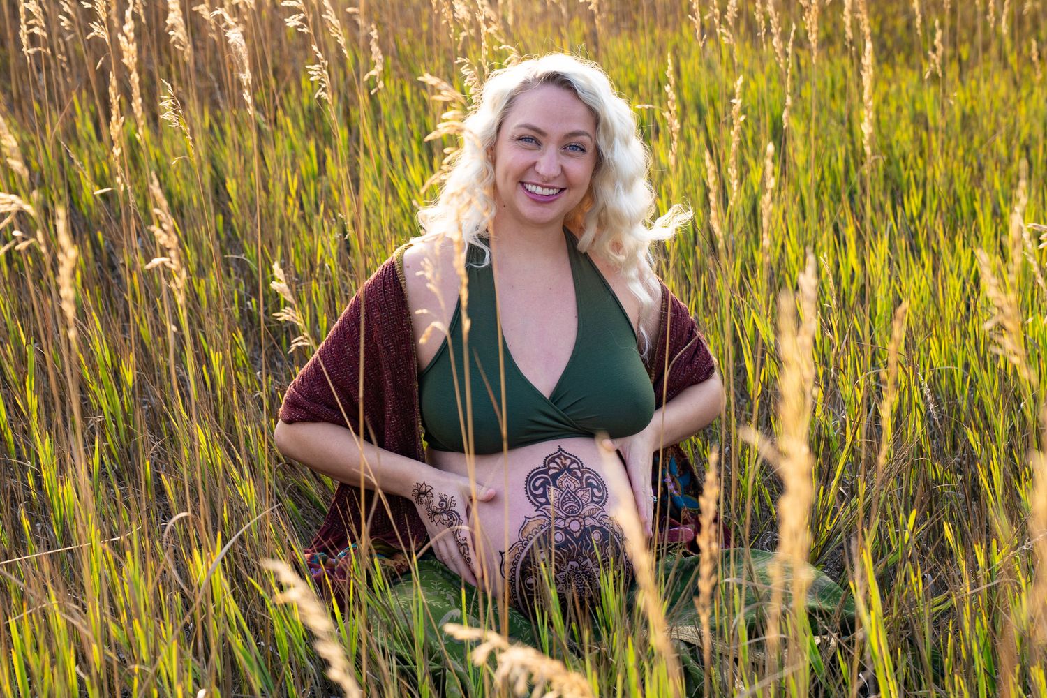 Smiling pregnant woman in green dress sitting in tall golden grass field during sunset maternity photo session.