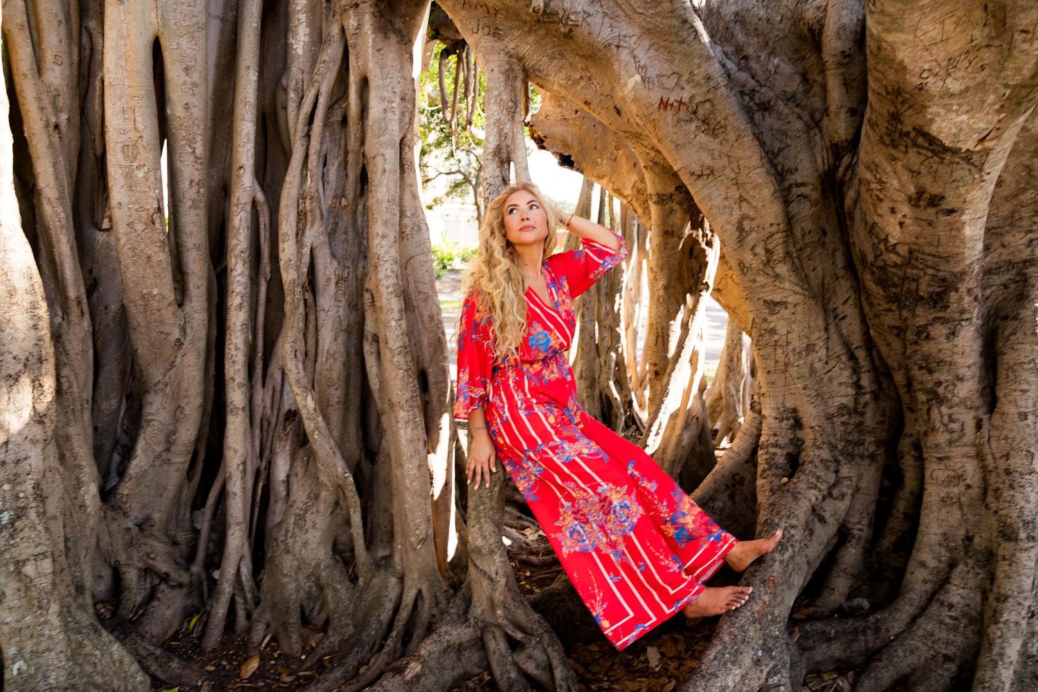 Woman in red floral maxi dress poses gracefully among massive twisted banyan tree roots in a serene outdoor setting.