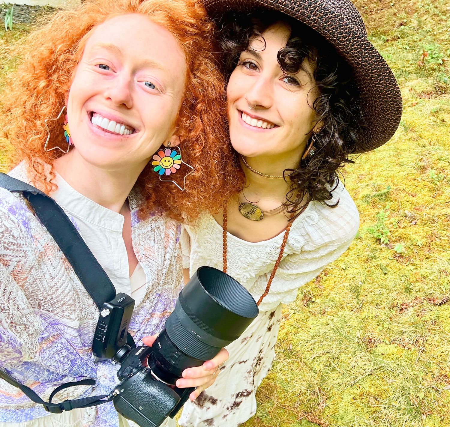 Two smiling women pose outdoors, one with red curly hair and flower earrings, the other in a hat holding a camera.