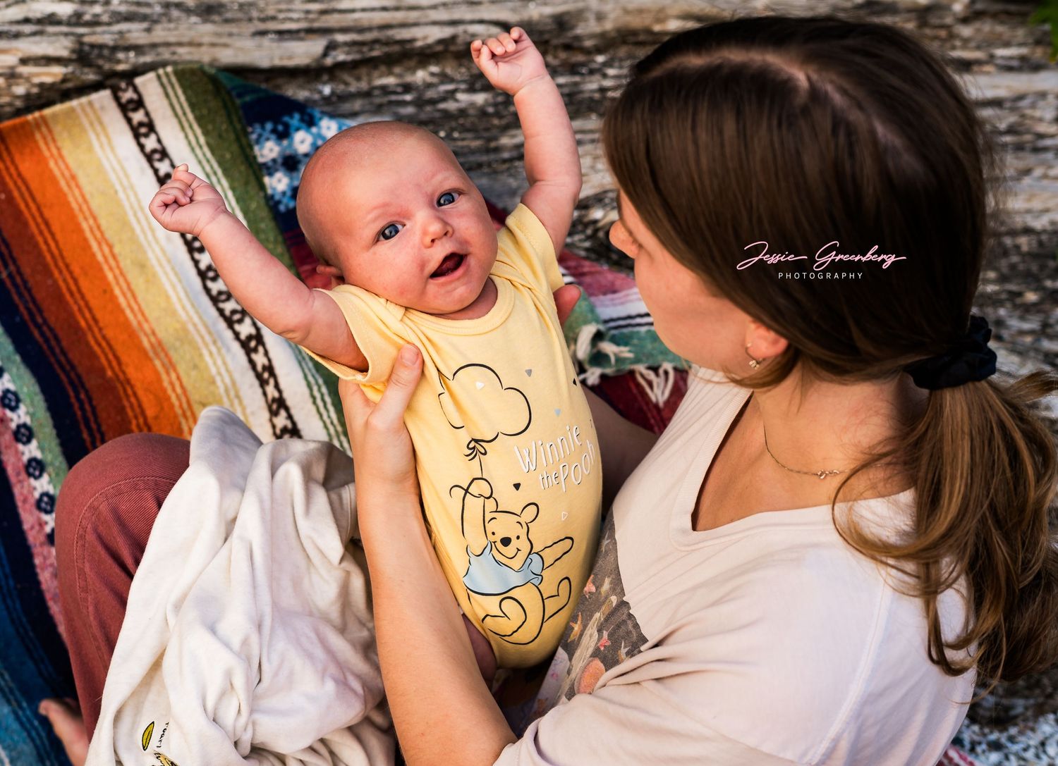 A mother lovingly holds her smiling baby dressed in a yellow Winnie the Pooh onesie during an outdoor photo session.