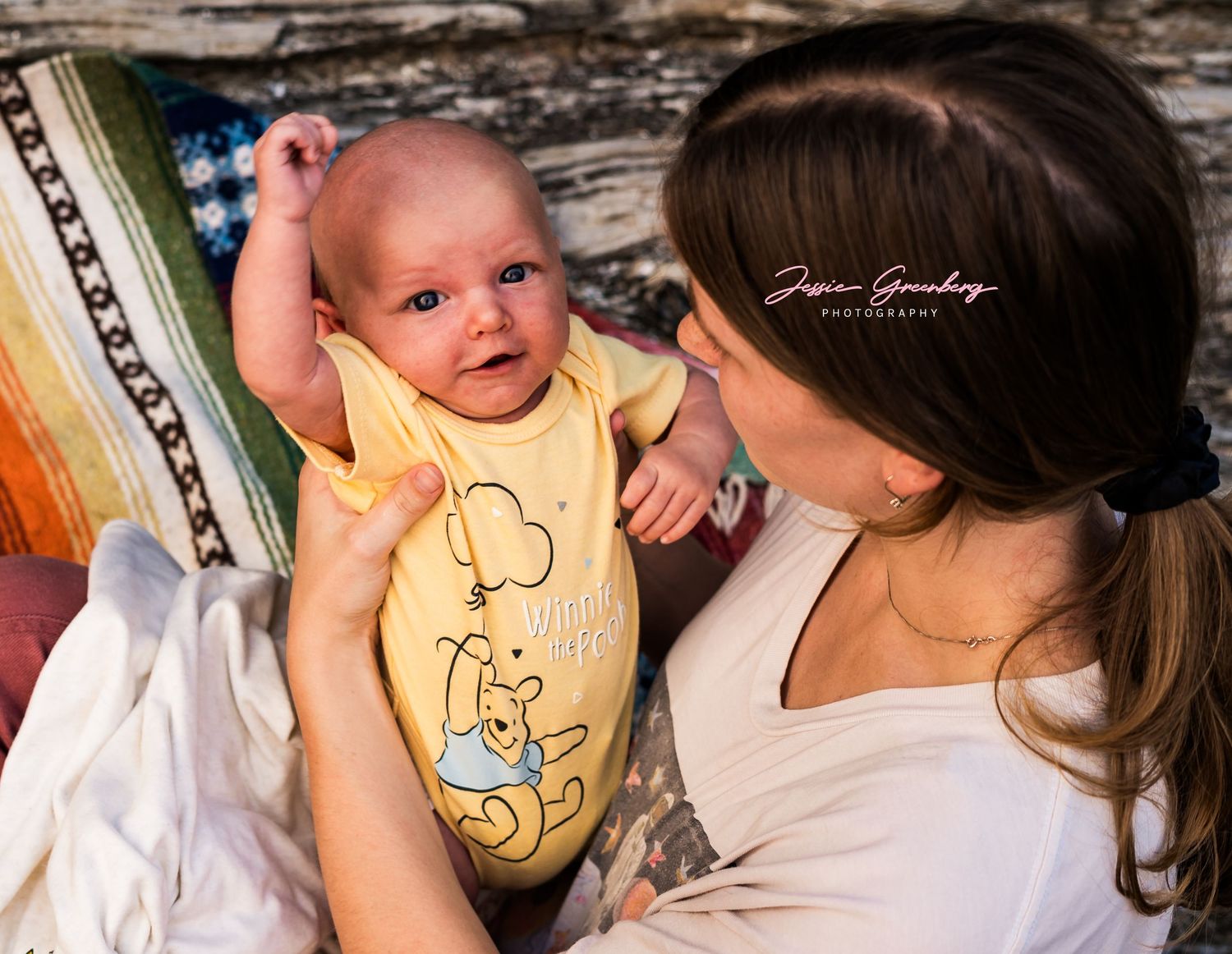 Mother lovingly holds smiling newborn baby wearing yellow Winnie the Pooh onesie during professional photography session.