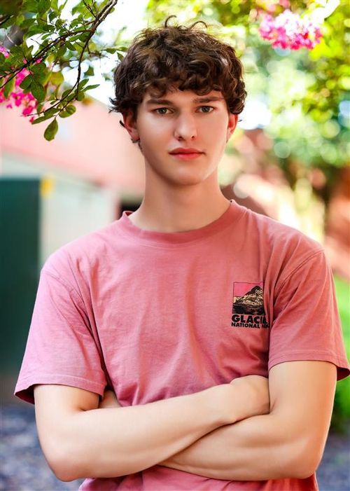 Young man with curly hair standing outdoors with arms crossed wearing a pink Glacier National Park t-shirt.