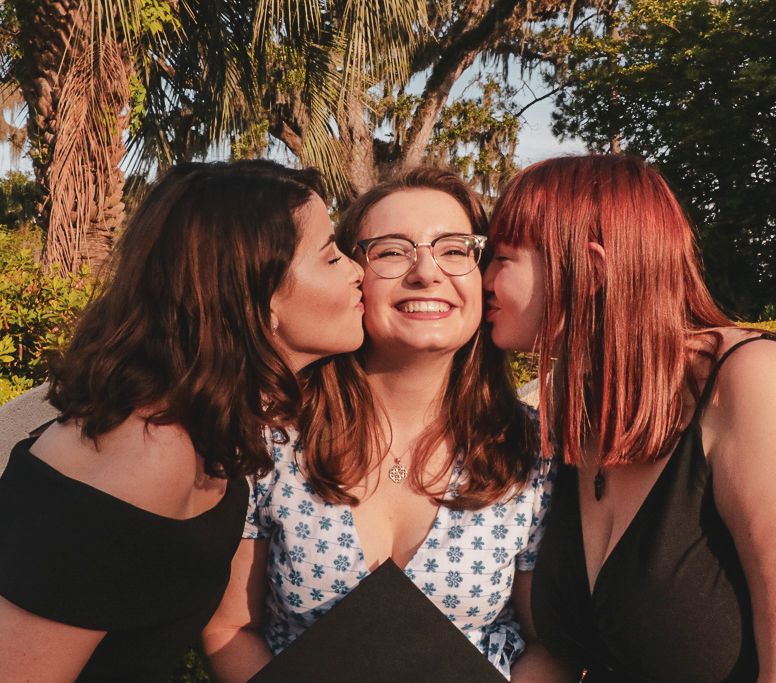 Three young women with auburn hair smile outdoors, two kissing the cheeks of the smiling middle woman wearing glasses.