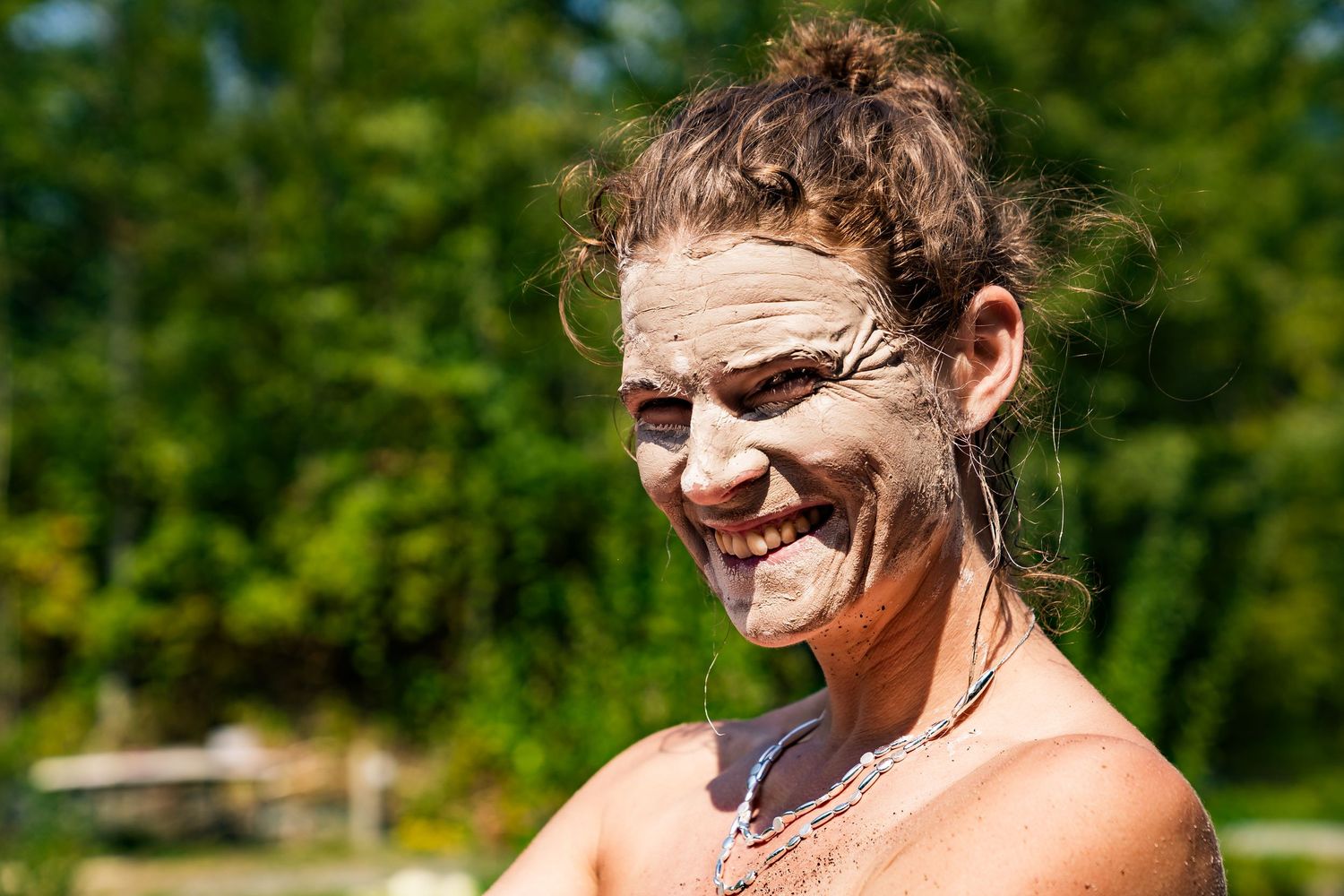 Smiling woman with mud mask on face outdoors, enjoying a natural mud treatment with green trees in background.