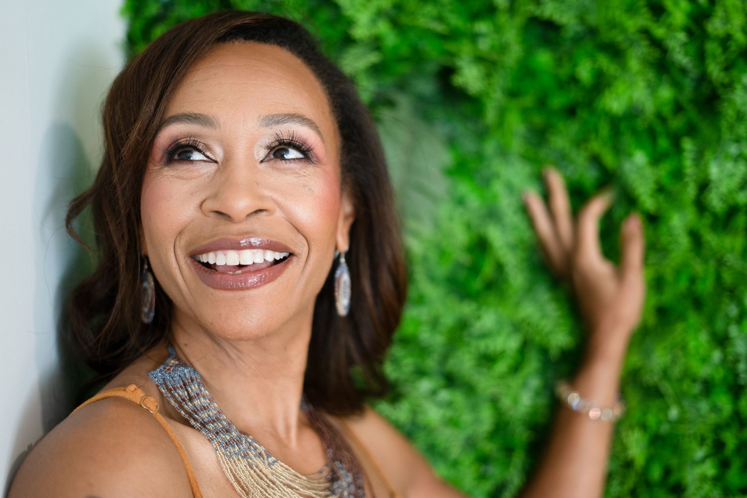 Smiling woman with diamond earrings laughing joyfully against a lush green plant wall backdrop.