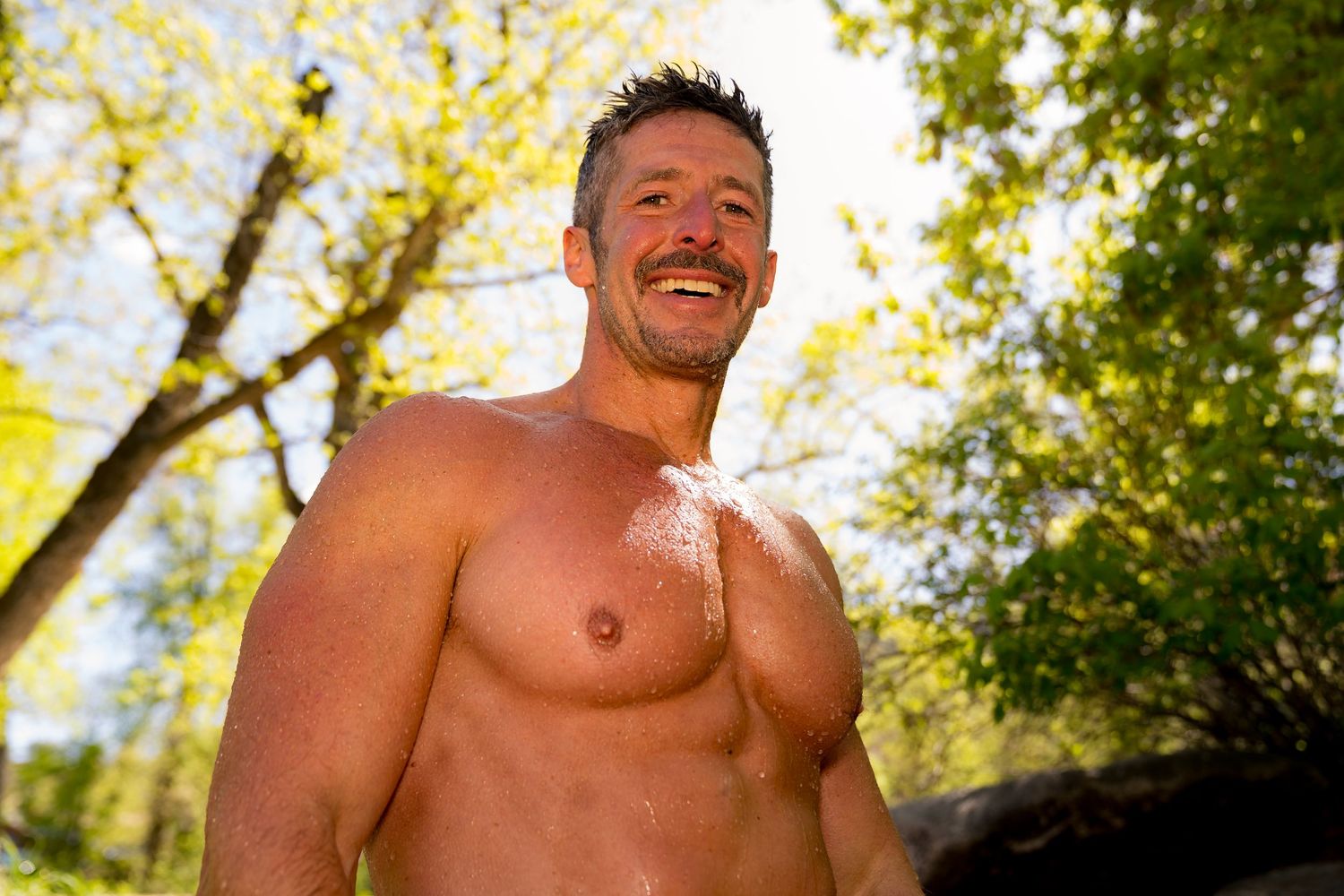 Muscular shirtless man smiling outdoors surrounded by green trees in natural sunlight.
