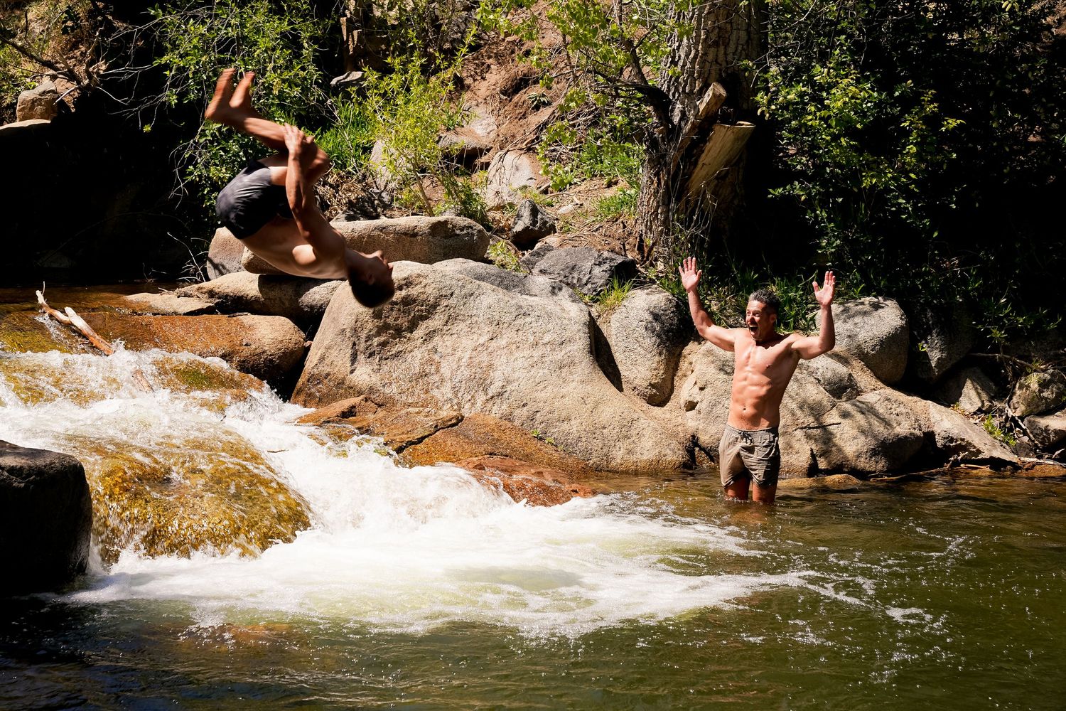 Two men jumping and playing in a rocky river waterfall surrounded by trees on a sunny day.