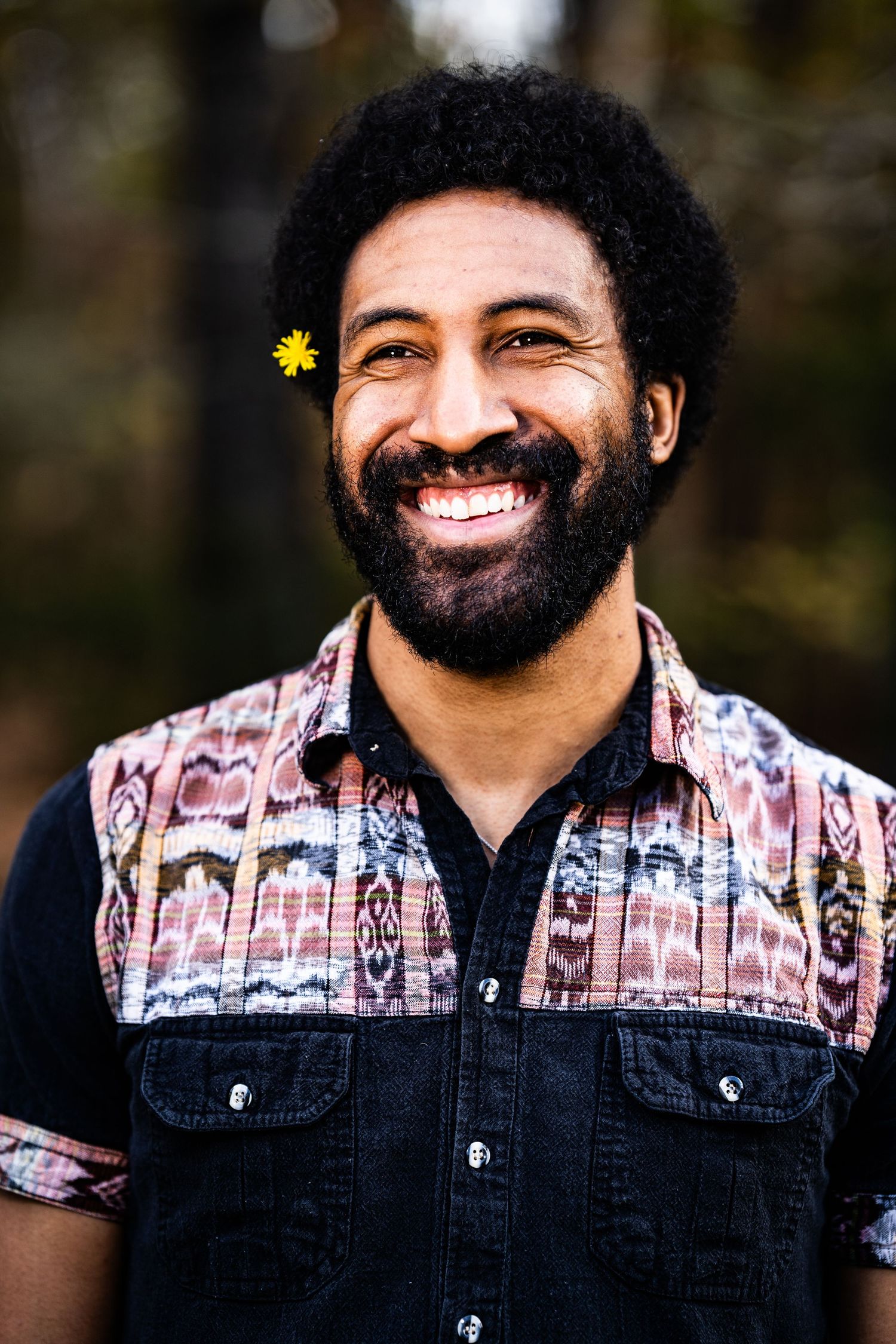 Smiling Black man with afro and beard, wearing a colorful patterned vest, with a small yellow flower tucked behind his ear.