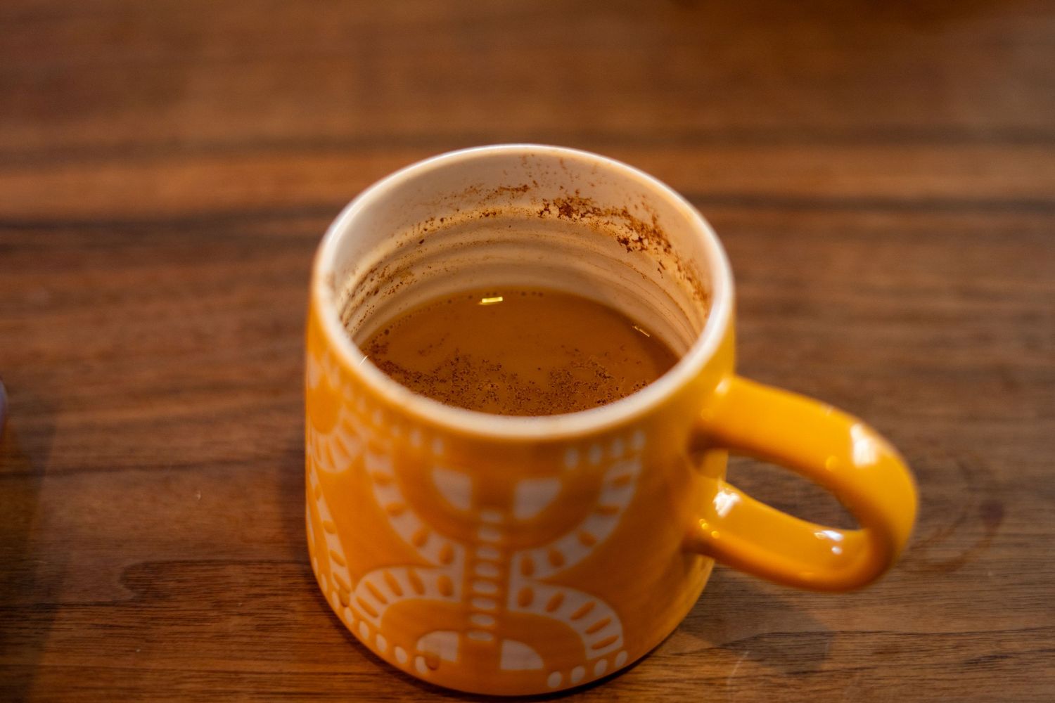 Orange ceramic mug with white patterns filled with hot chocolate topped with cinnamon on a wooden table.