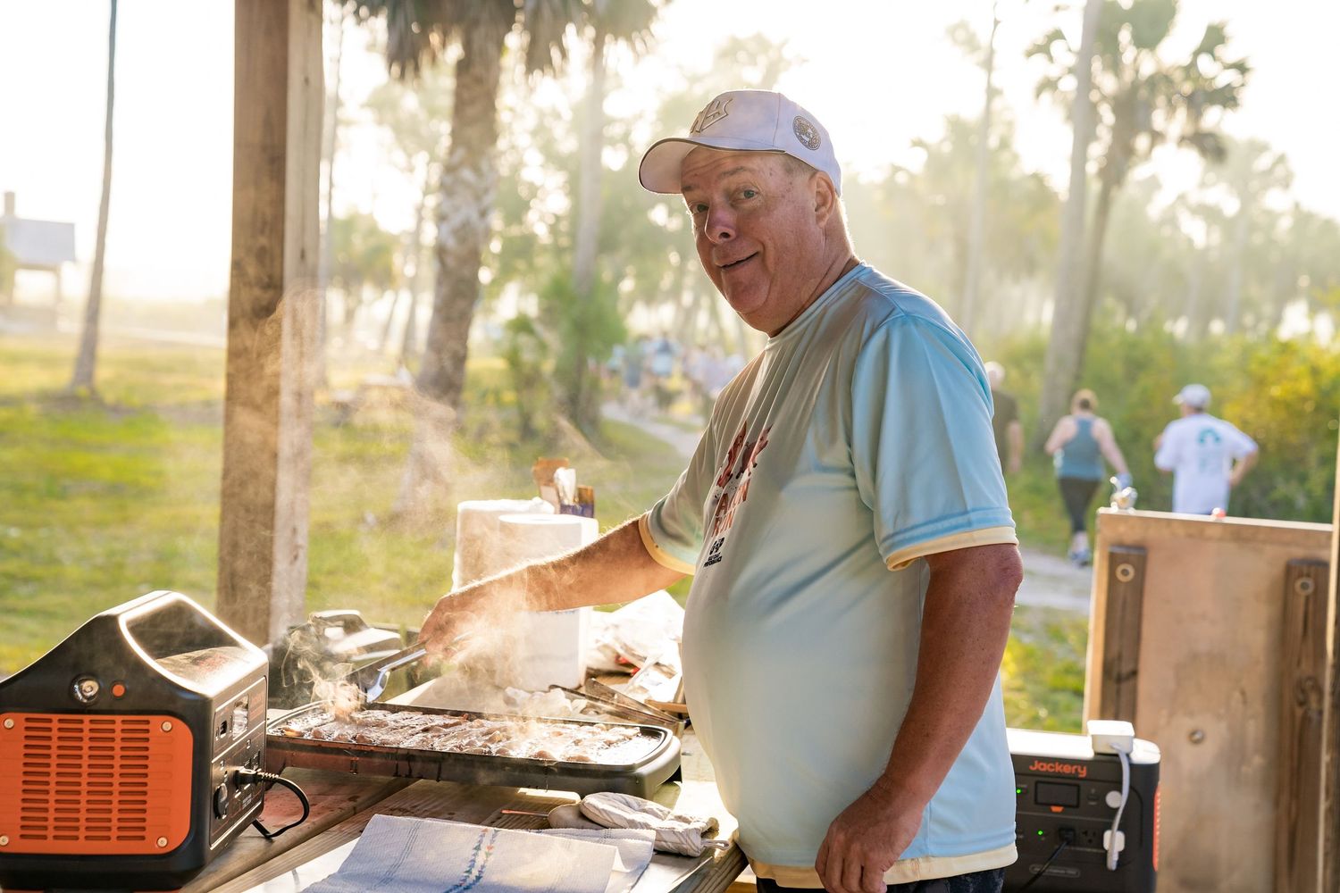 A man using a grill, looking at the camera as smoke rises around him.