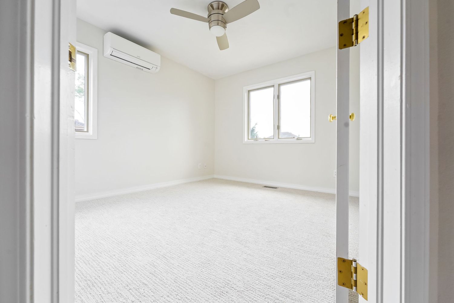 Empty white bedroom with ceiling fan, wall-mounted AC unit, carpet flooring, and two windows providing natural light.