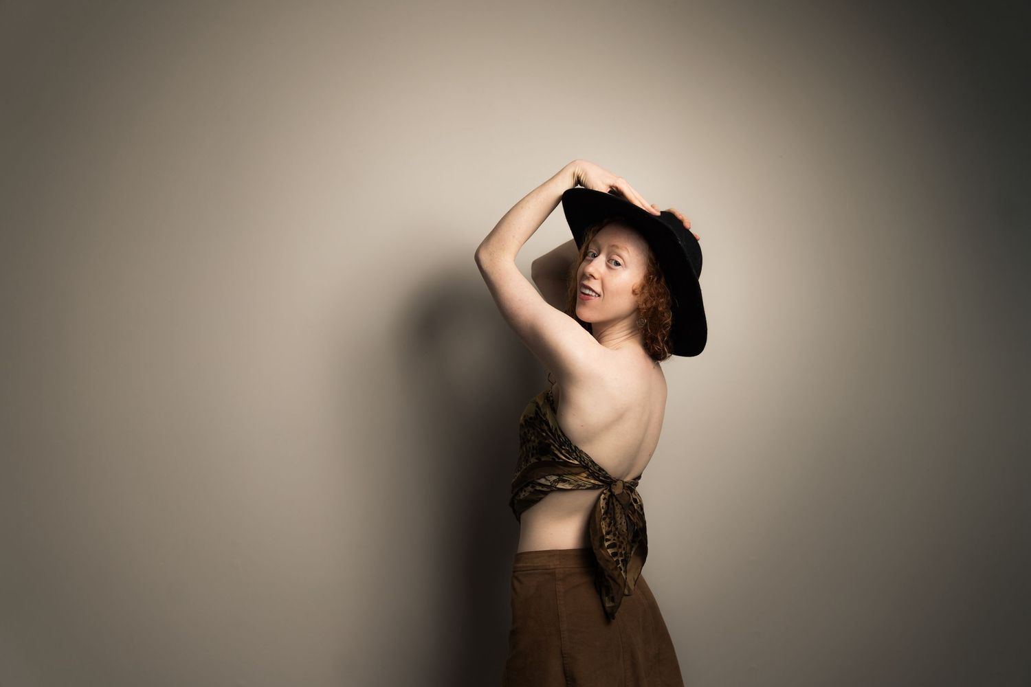 Woman in black hat and backless brown outfit posing gracefully against a neutral gray background.