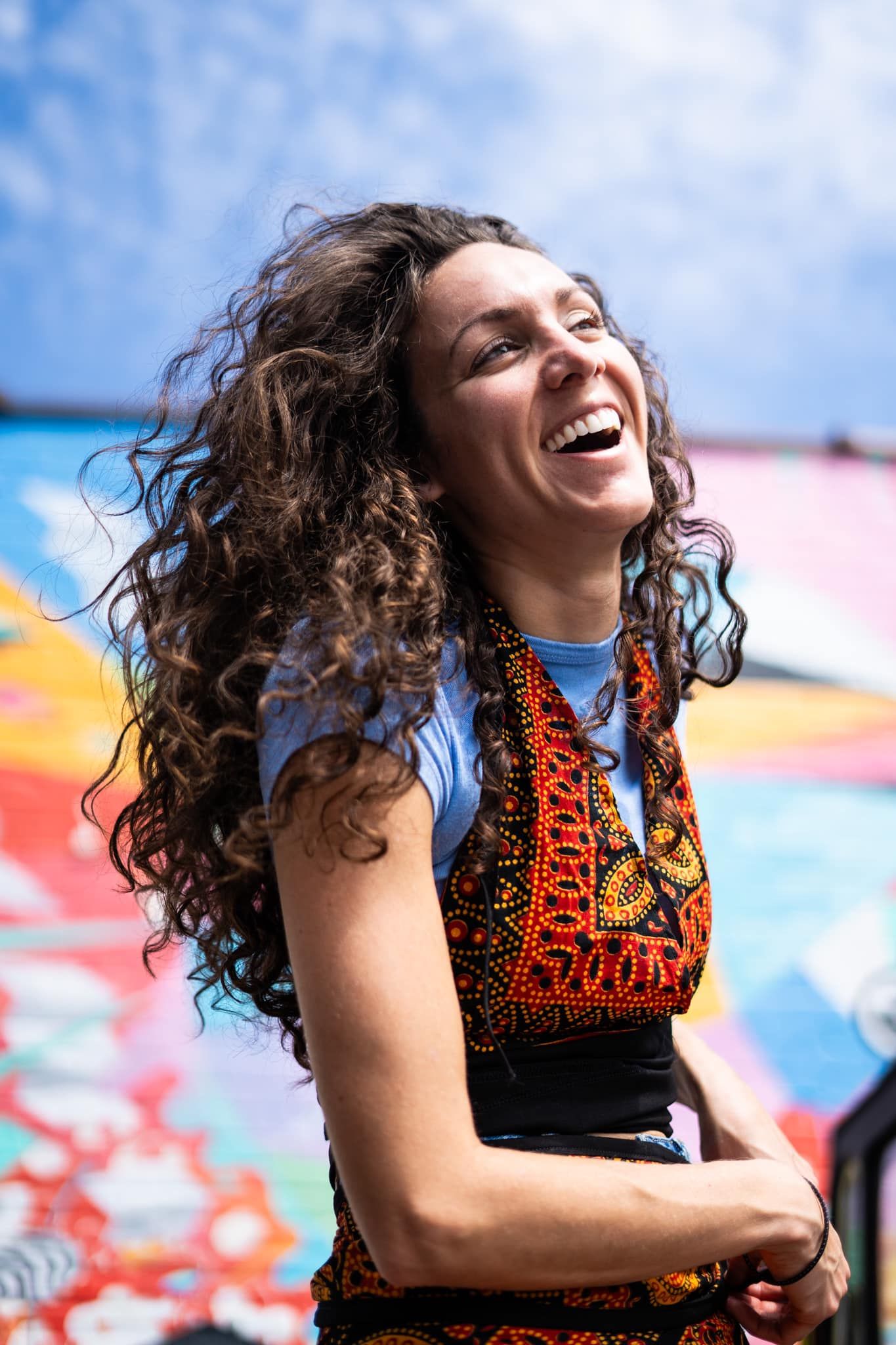 Laughing woman with curly hair wearing colorful ethnic vest stands in front of vibrant street art mural outdoors.