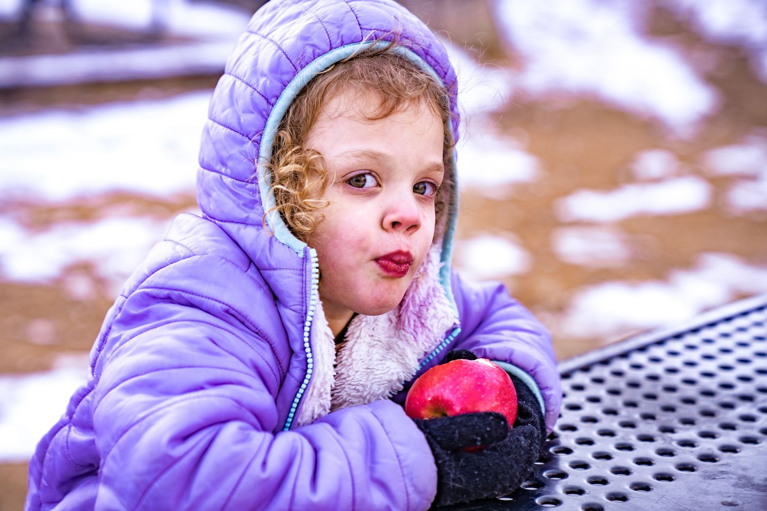 A small child in a purple winter coat leans against a metal railing while playing outdoors in snowy weather.