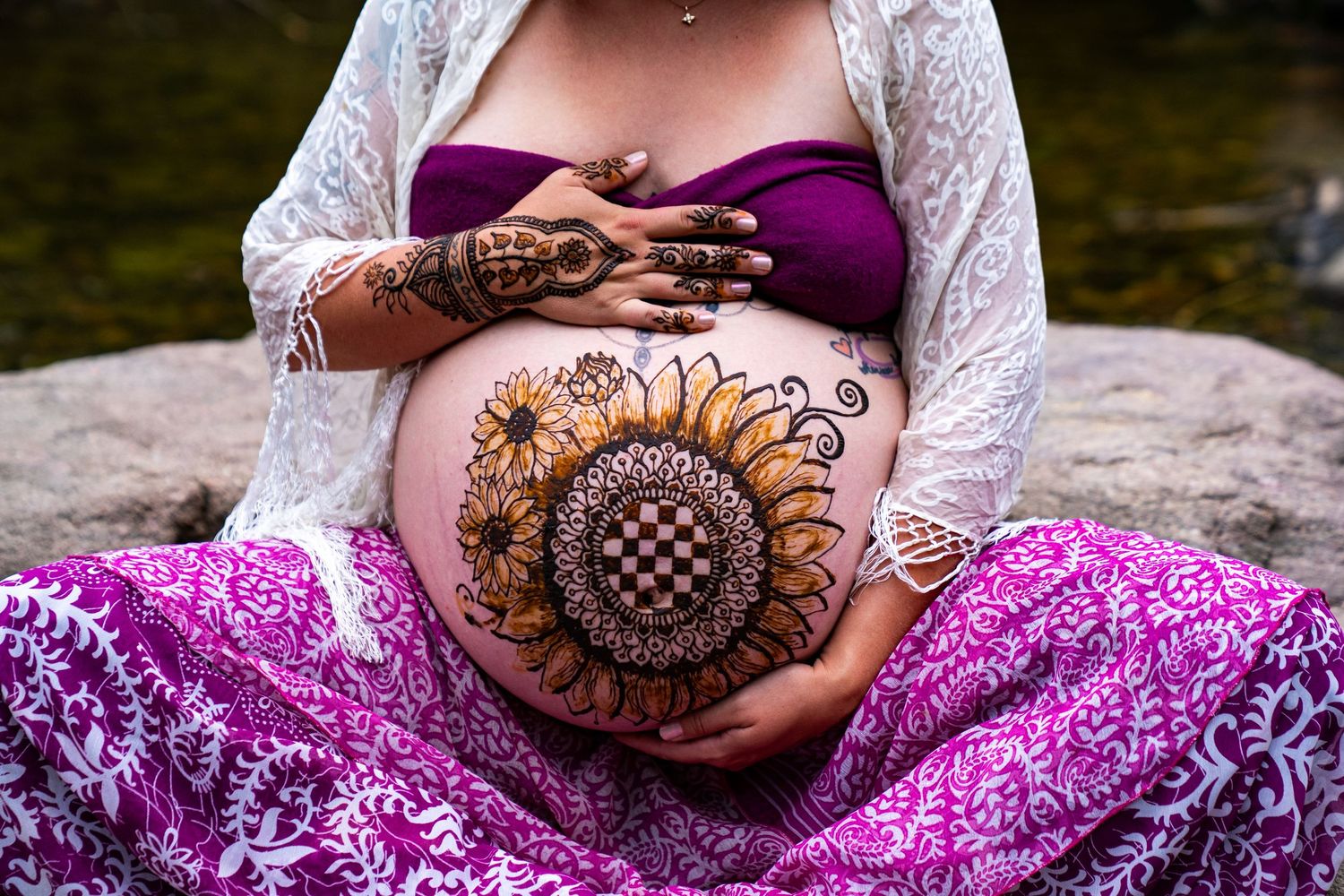 Beautiful henna art featuring a large sunflower design decorates a pregnant belly against a purple skirt background.