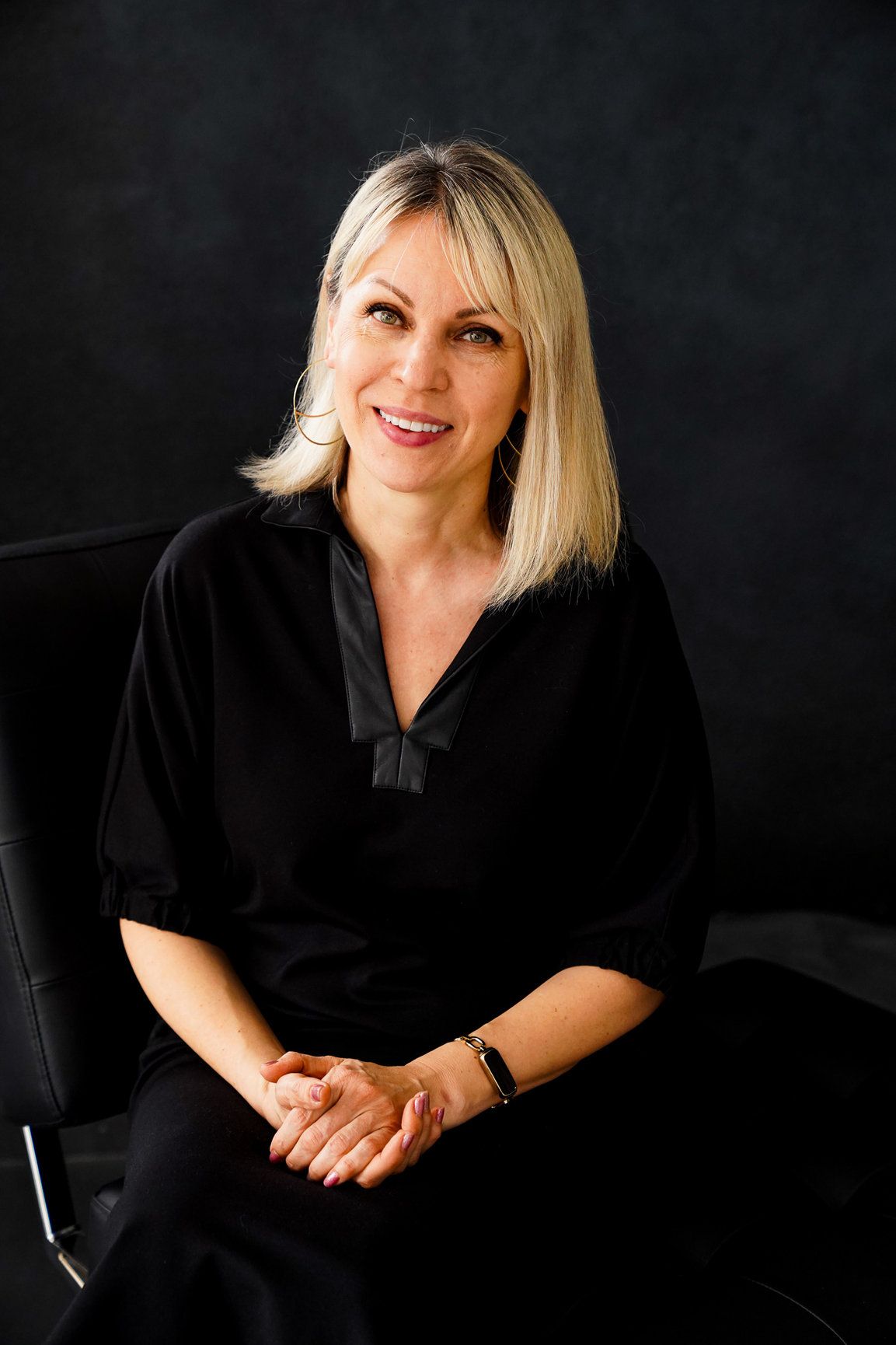 A woman wearing black in a studio with a black backdrop smiling and looking at the camera.
