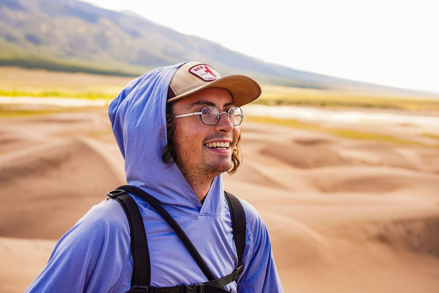 A photo of a guy with a blue jacket and a hood he is smiling off to the side with glasses on.