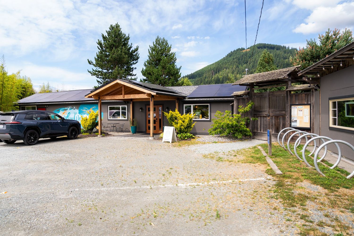 Solar-paneled commercial building with bike rack, gravel parking lot, and mountain backdrop in a Pacific Northwest setting.
