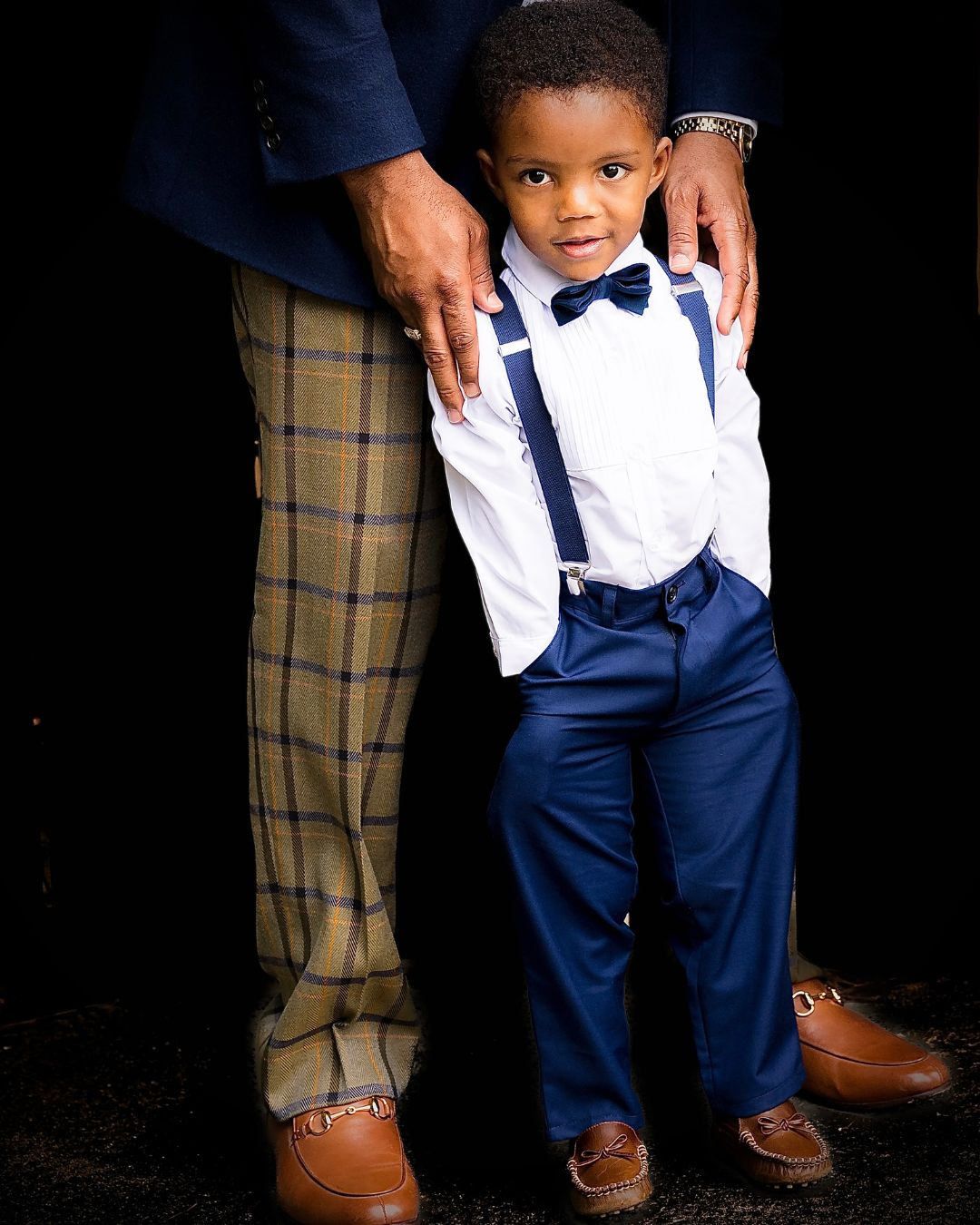 Adorable young boy in formal navy suit with suspenders and bow tie standing in front of an adult outdoors.