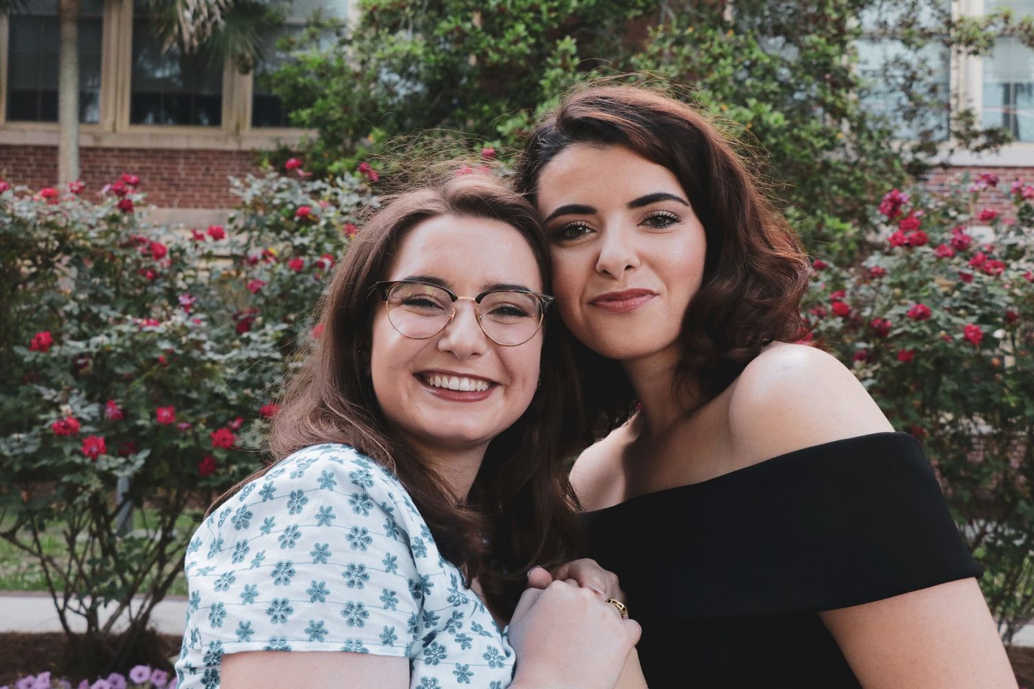 Two young women smiling and posing together outdoors near blooming red flowers and green trees.