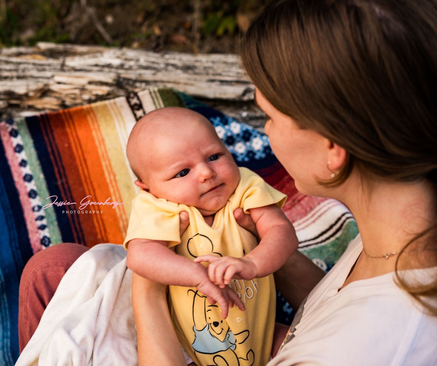 A mother tenderly holds her smiling newborn baby dressed in a yellow Winnie the Pooh onesie outdoors.