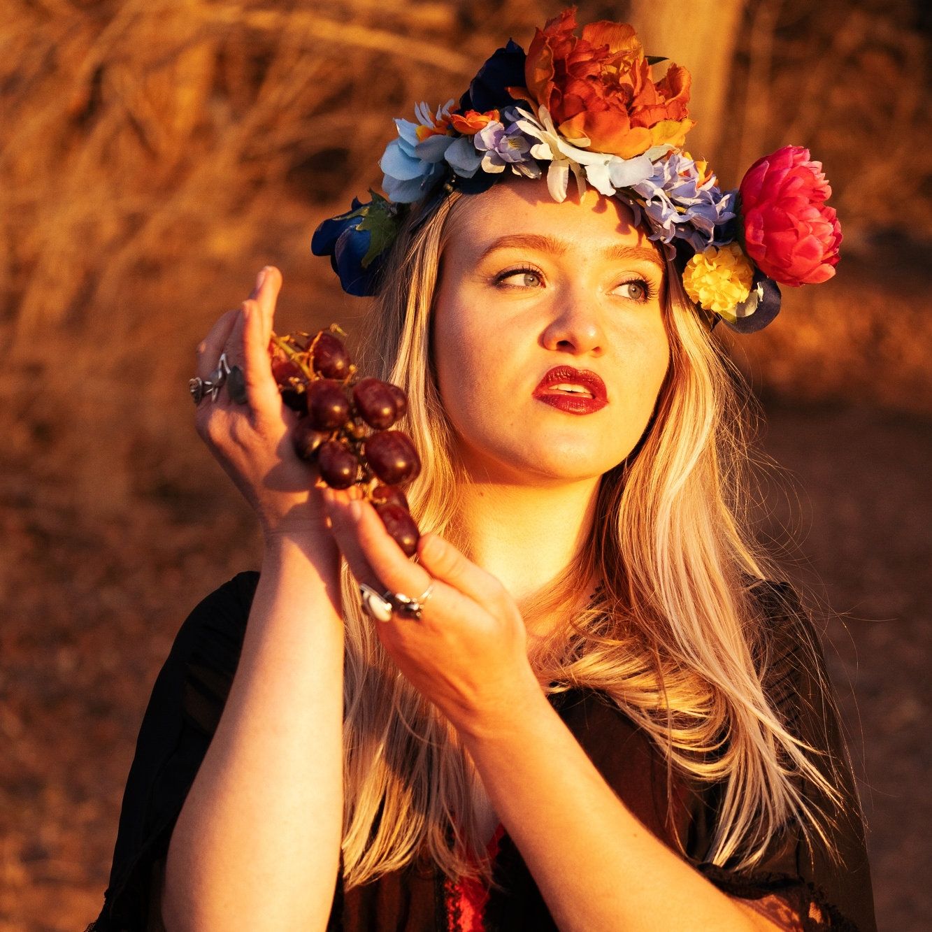 A photo of a woman wearing a flower crown and holding red grapes, she has blonde hair and a black top.