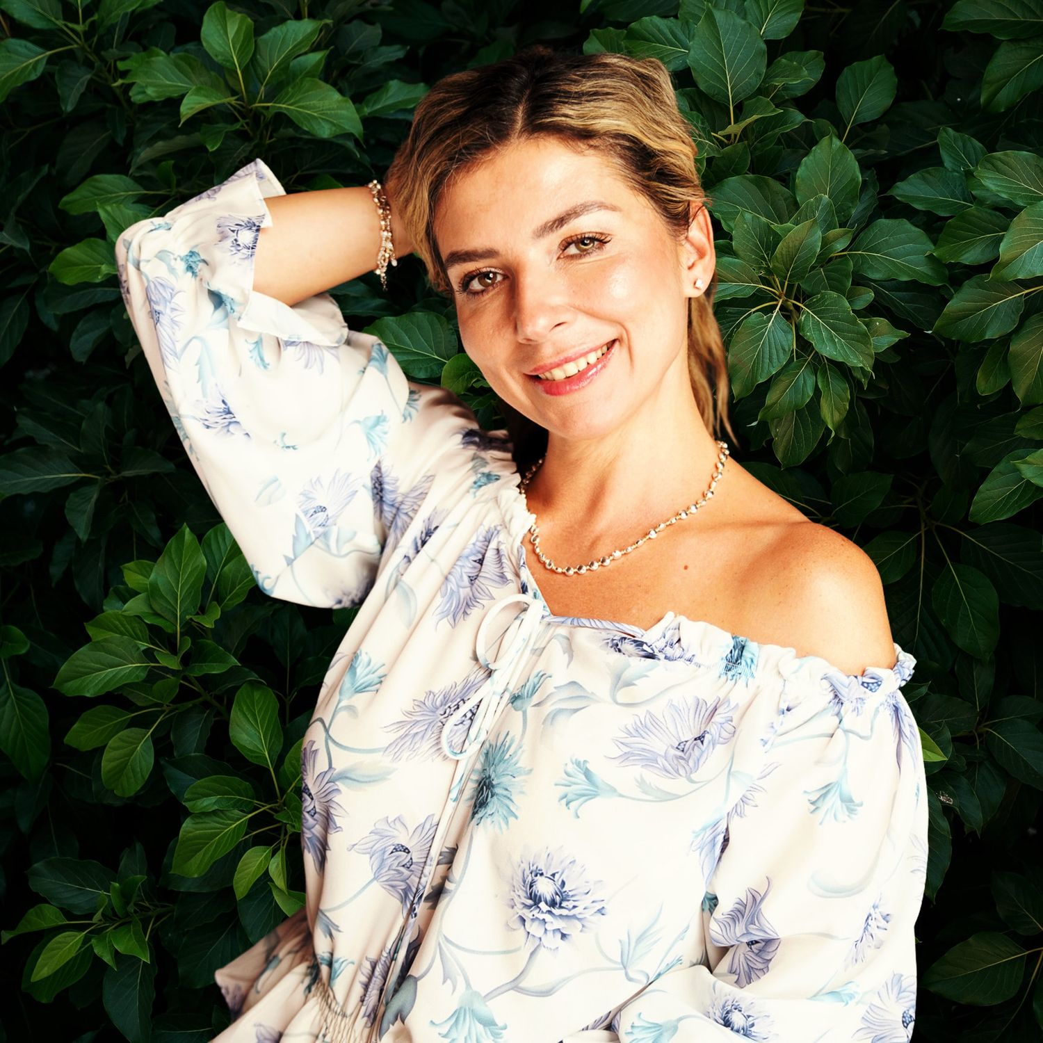 A photo of a woman with blonde hair and her shirt draped off her shoulder against a backdrop of green leaves.
