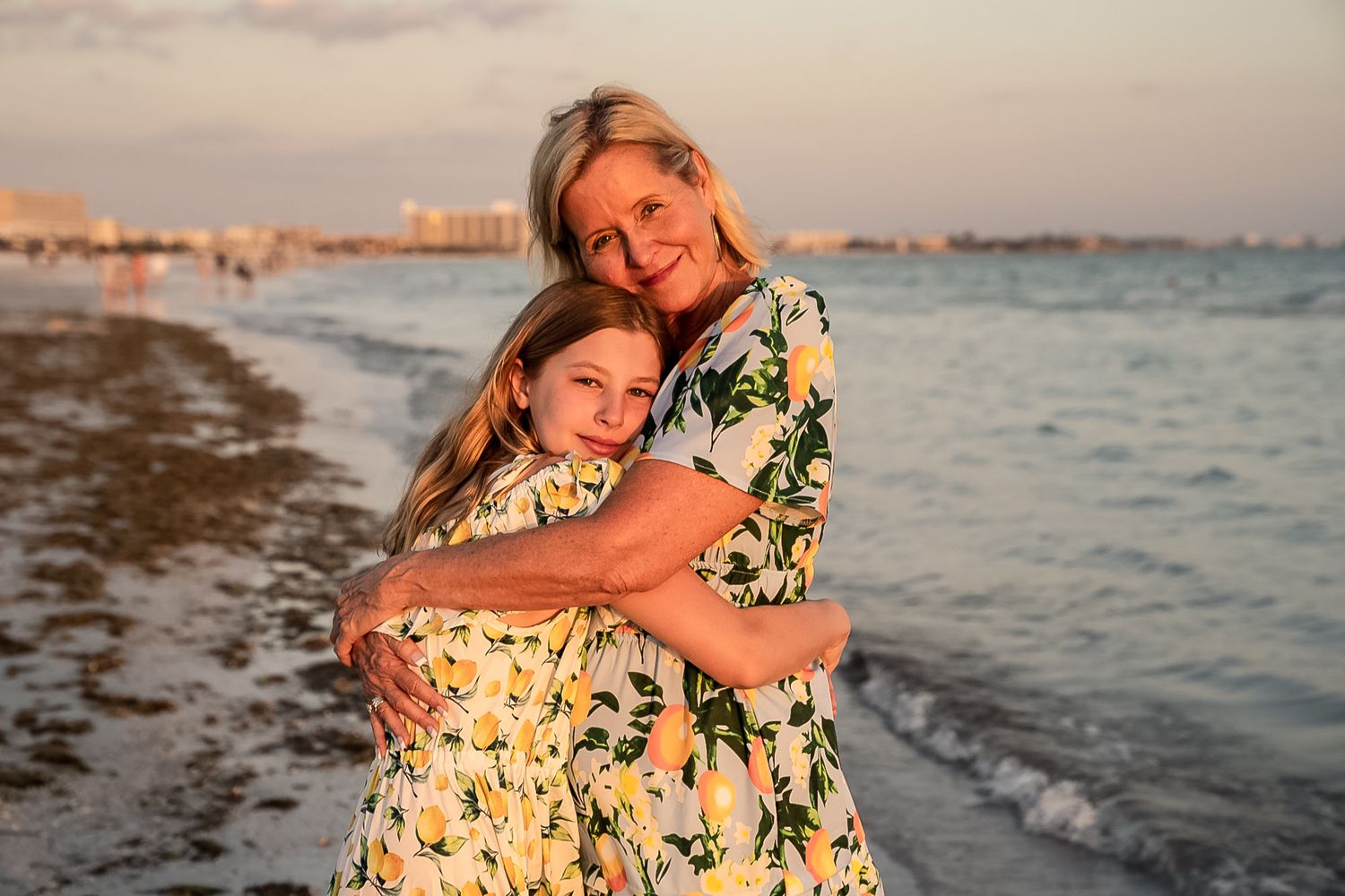 A photo of a mother and a daughter hugging with water and seaweed behind them with a sunset.