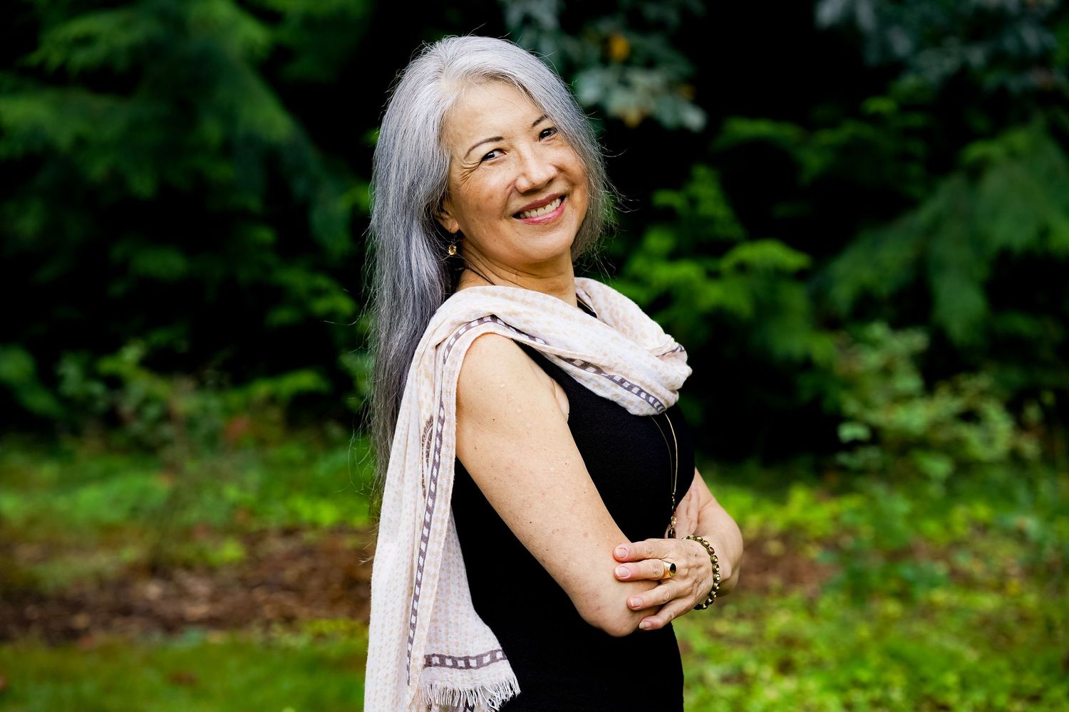 Smiling older woman with long gray hair wearing black sleeveless top and white scarf, standing outdoors in lush green garden.