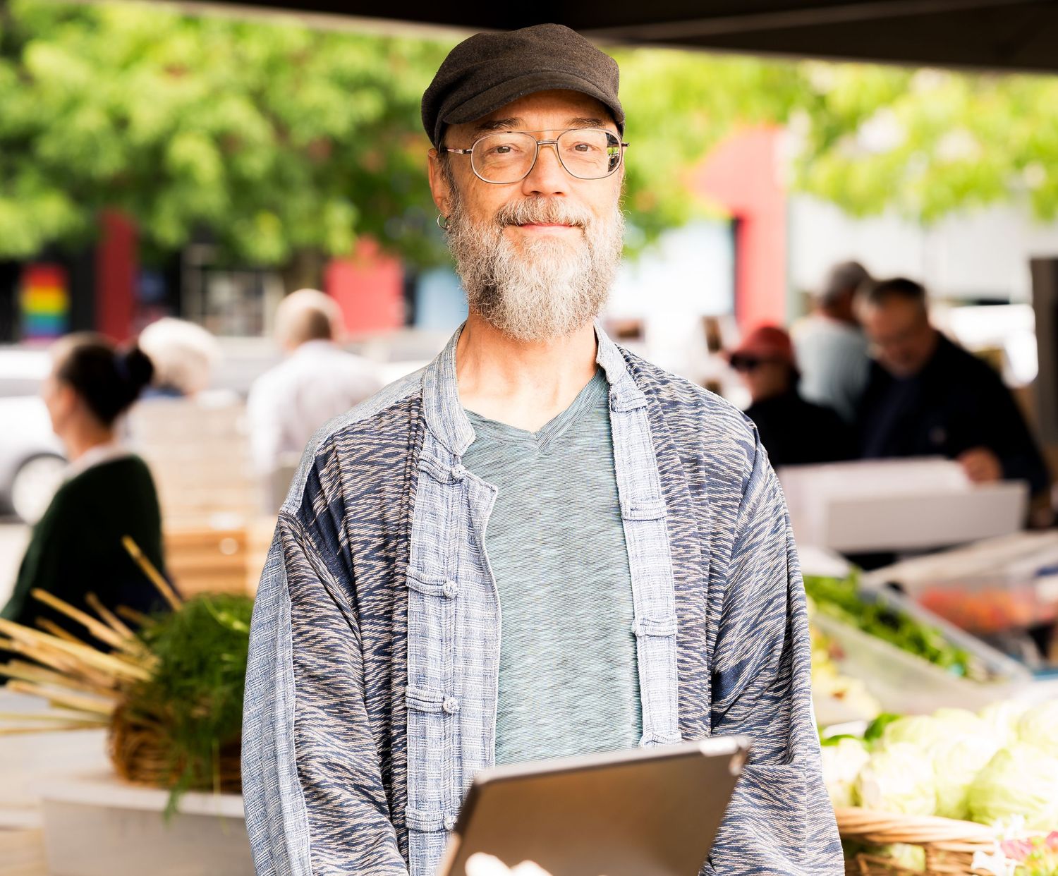 Smiling older man with beard and cap holding tablet at outdoor farmers market with fresh vegetables.