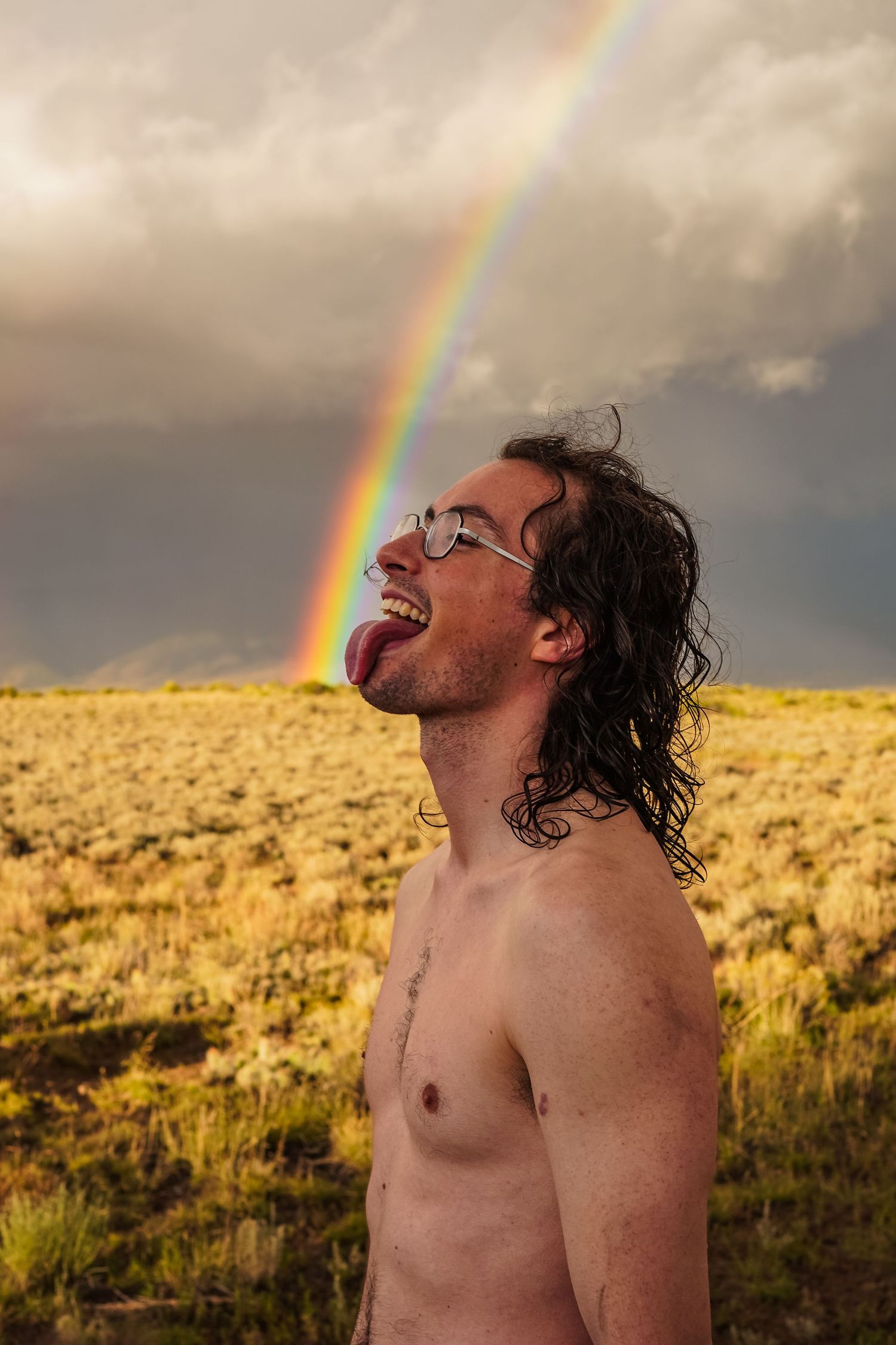 a photo of a guy with curly brown hair licking a rainbow, he is shirtless and had brown grass behind him.