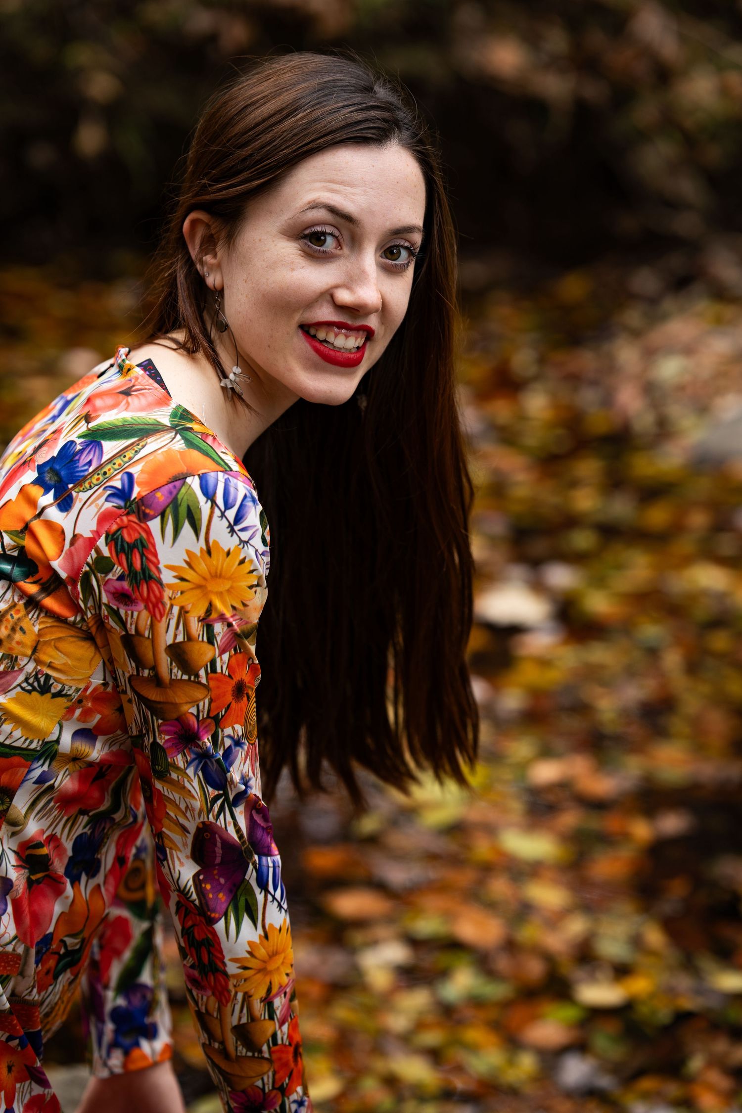Woman in colorful floral dress poses playfully in autumn forest setting with fallen leaves on ground.