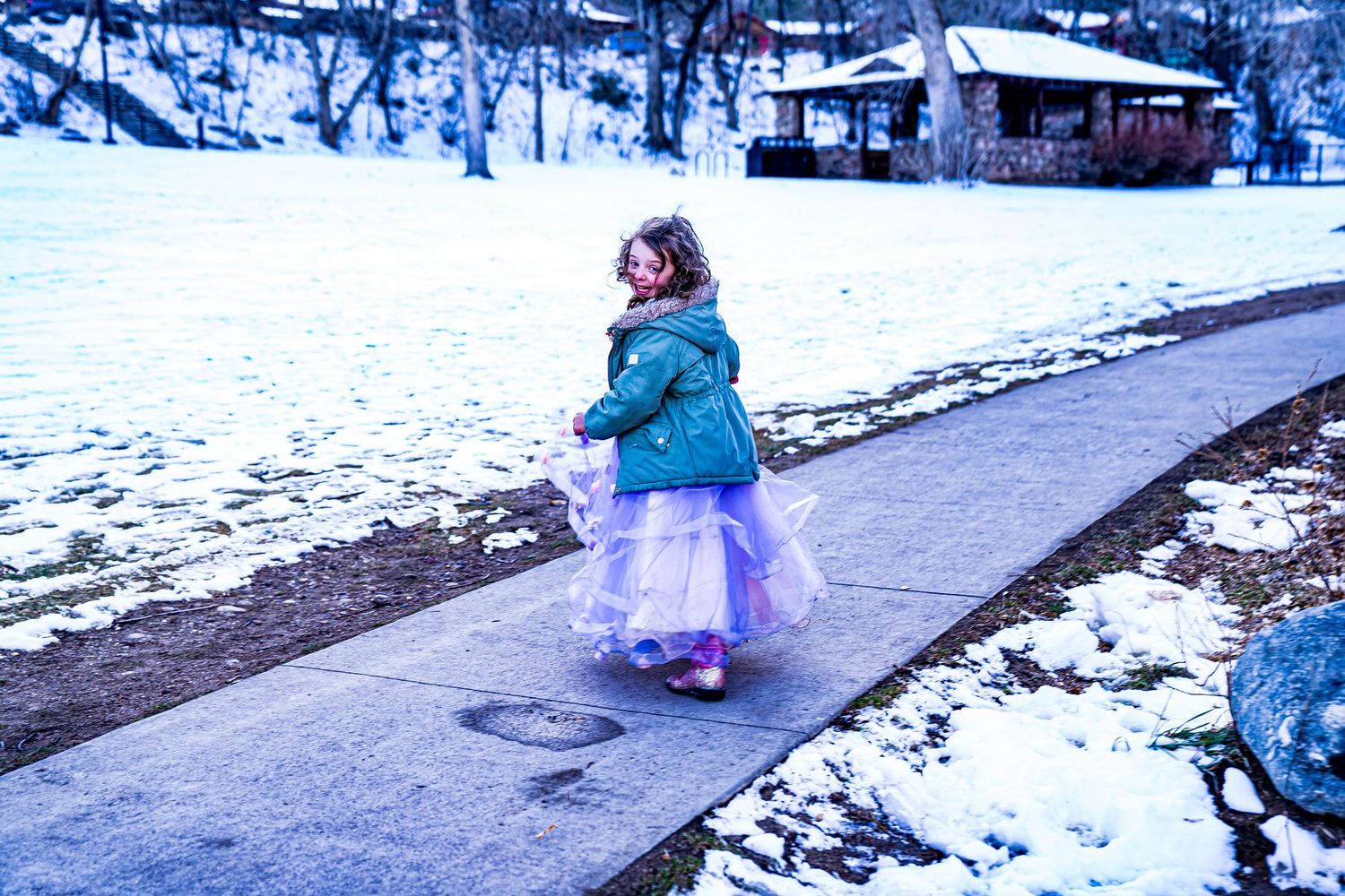 A child in a teal winter coat and tulle skirt walks along a snowy path with buildings visible in the background.