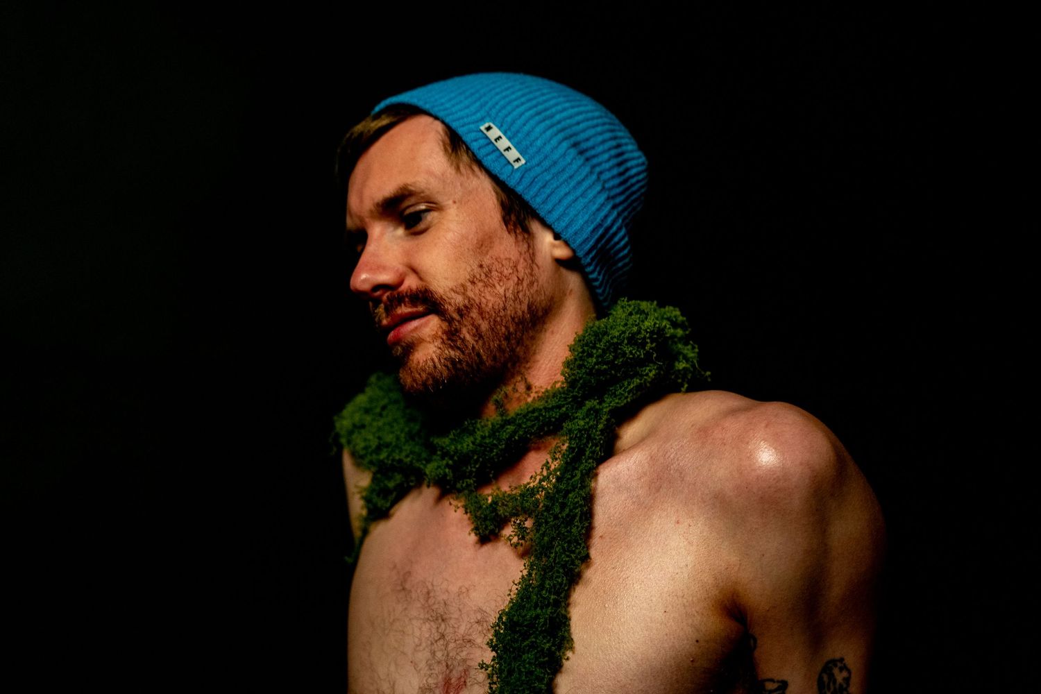 A shirtless person wearing a blue beanie and draped with green garland poses against a dark background.