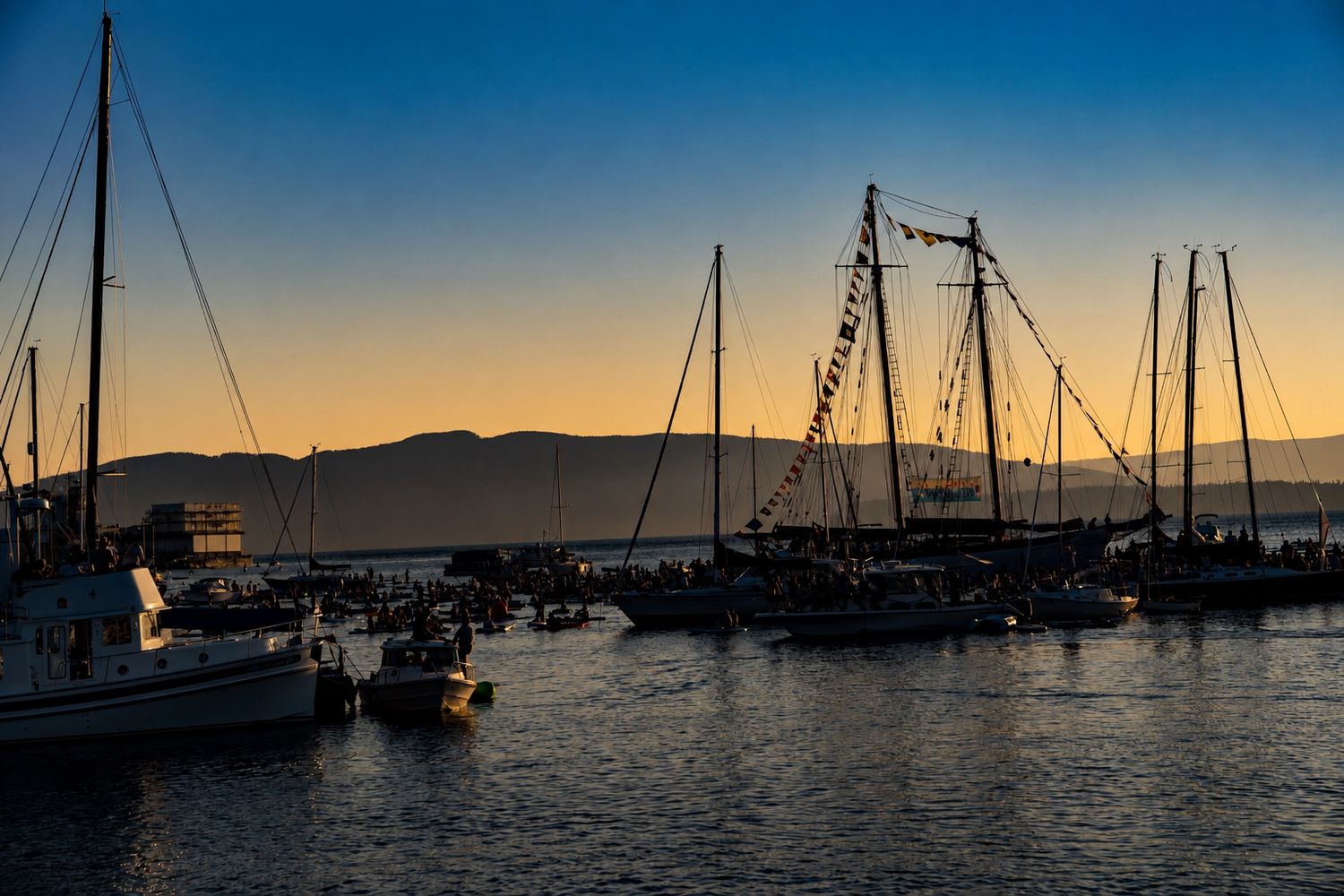Sailboats silhouetted against a golden sunset sky in a calm marina with mountains in the background.