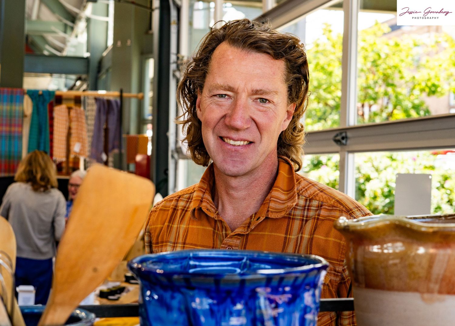 Smiling man with curly hair in orange flannel shirt at a craft market with blue ceramic pottery in foreground.