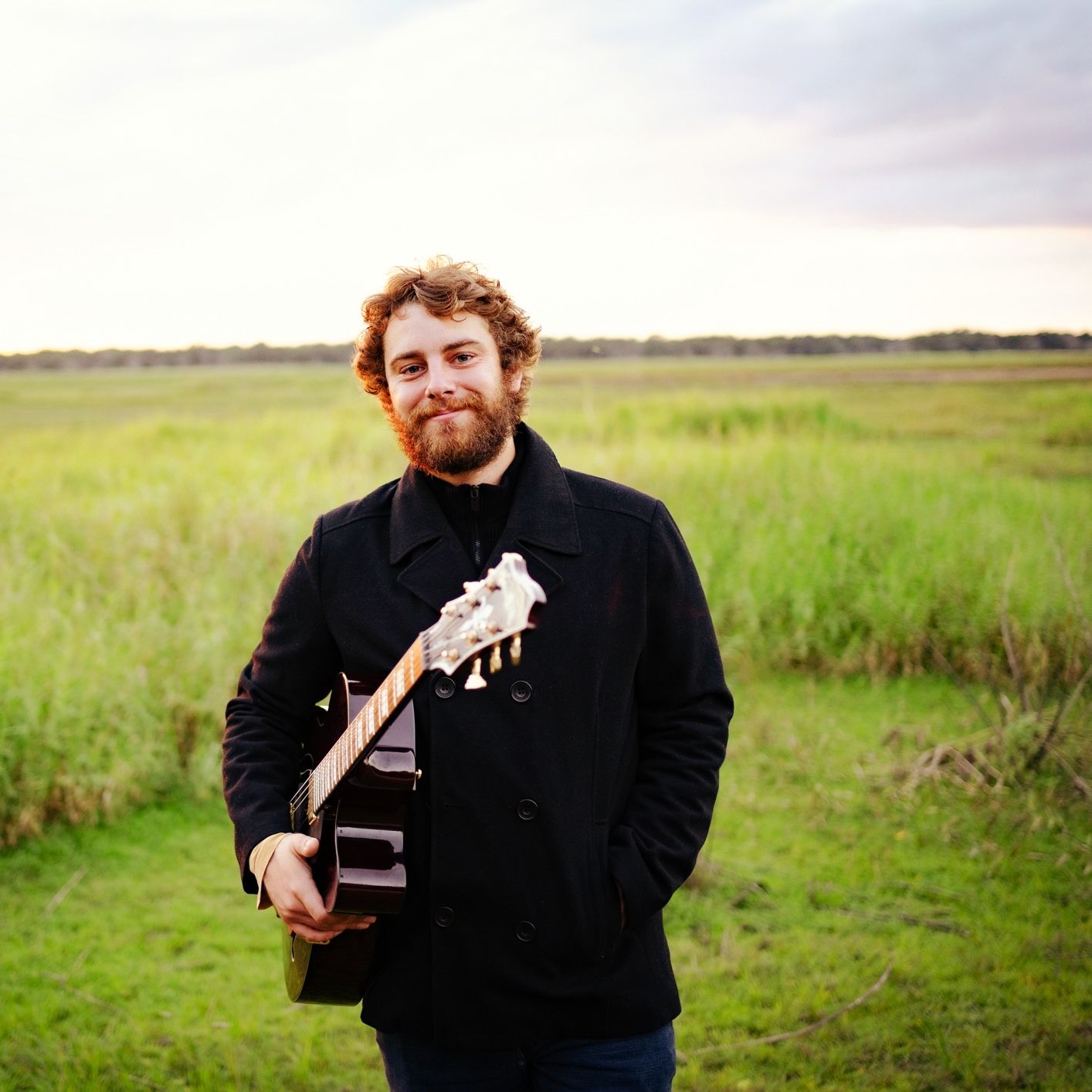 A bearded man with curly hair holds a guitar while standing in a green field under a cloudy sky.