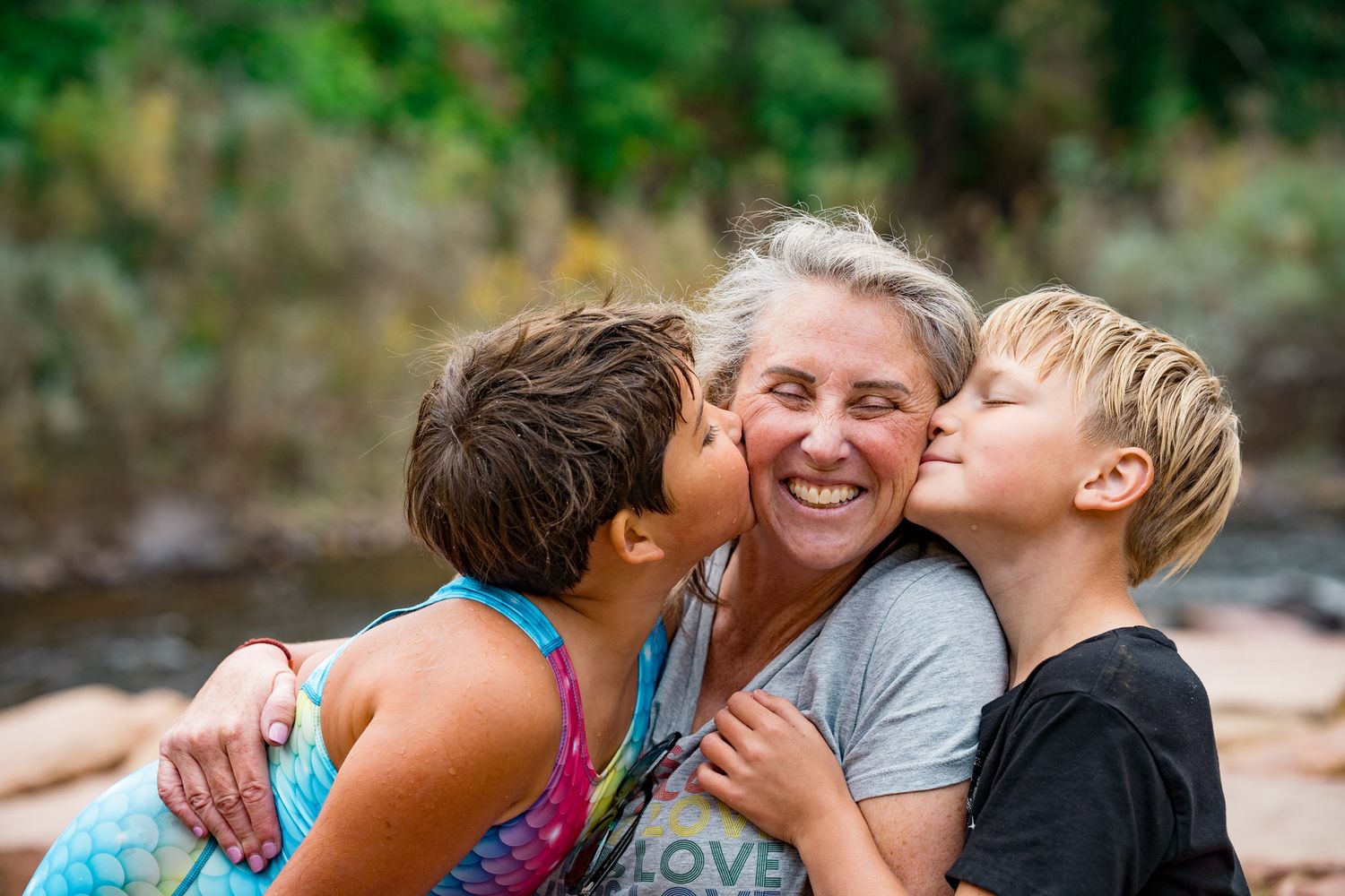 Two children kissing a laughing older woman on her cheeks outdoors near a river.