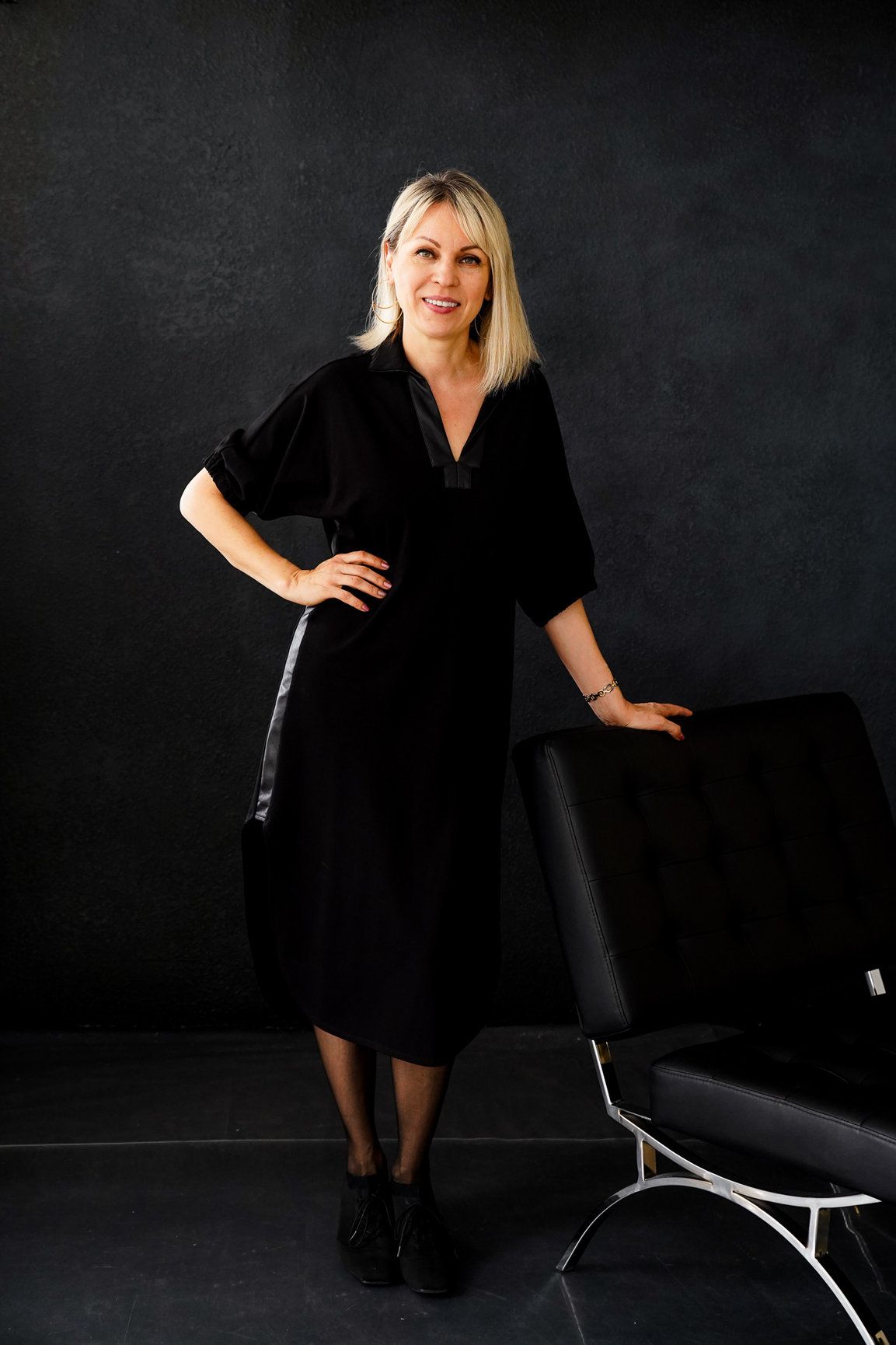 A photo of a woman with her hand on hip and one hand on the black chair with studio lighting.