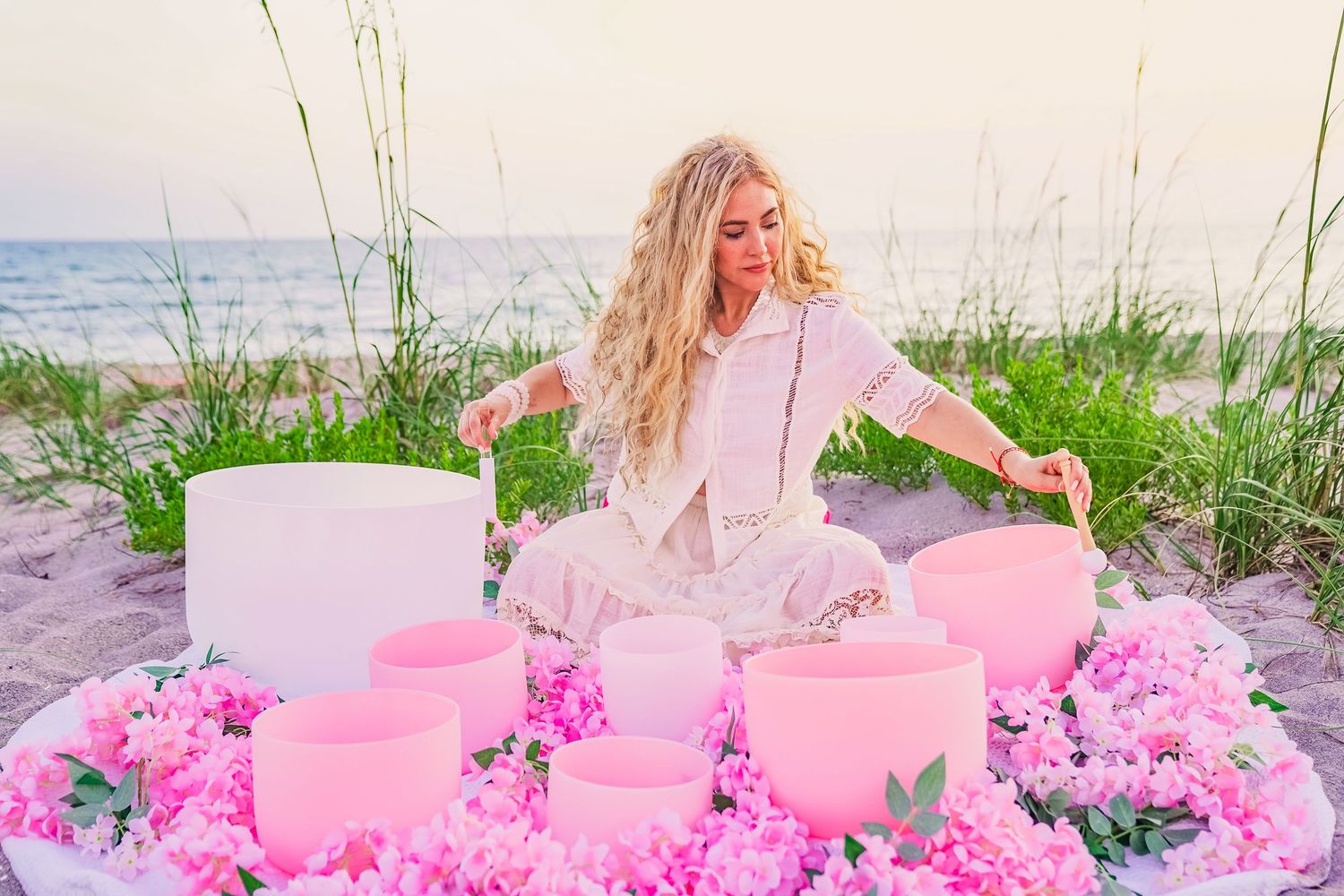 Dreamy pink beach scene with crystal singing bowls and scattered flowers on sandy shore at sunset.